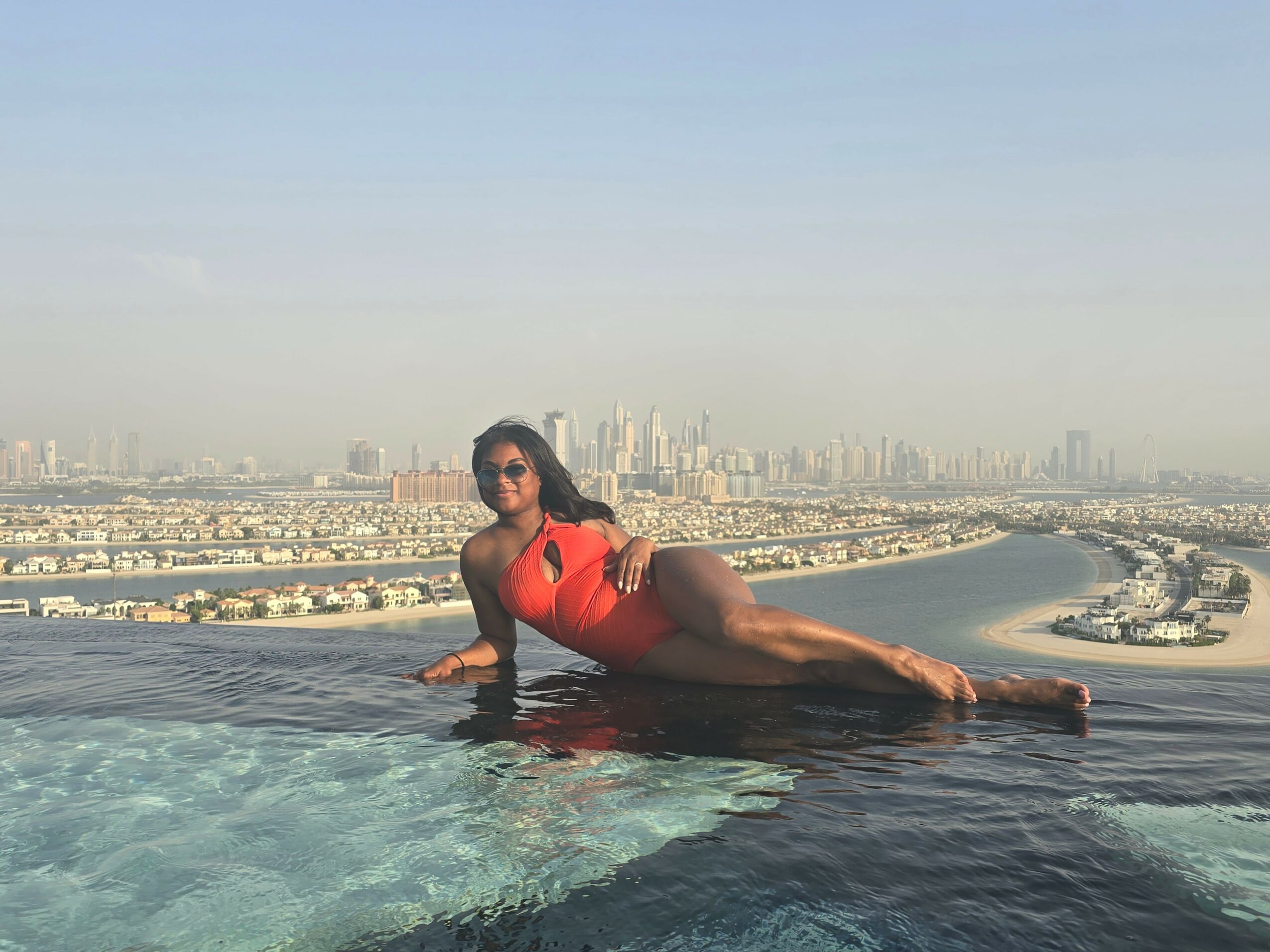 Woman in red swimsuit lying on edge of infinity pool with city skyline in background.