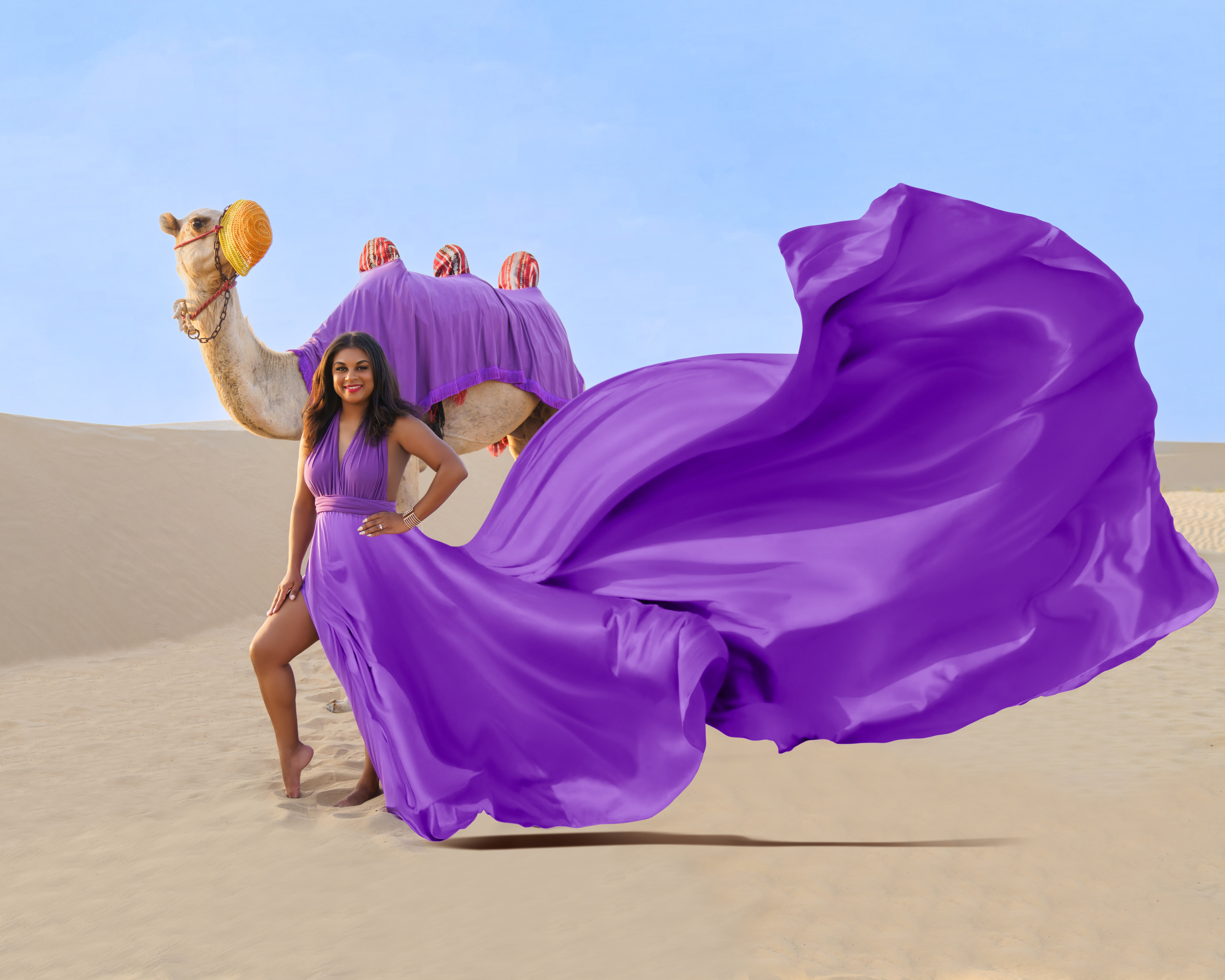 Woman in a purple dress standing on sand with a purple and beige camel costume behind her.