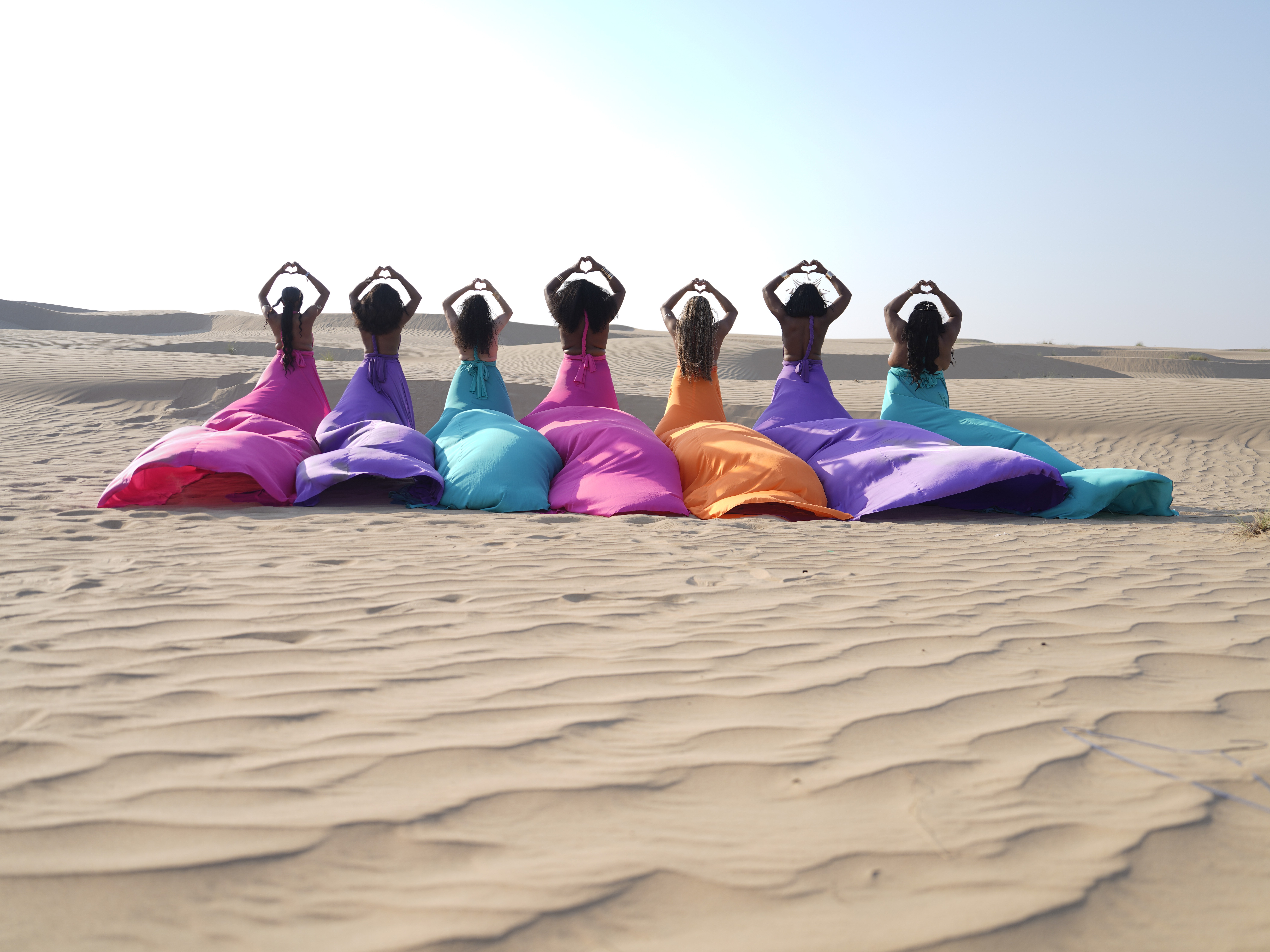Six women in colorful dresses sit on sand dunes with hands on heads, facing the camera in a desert landscape.