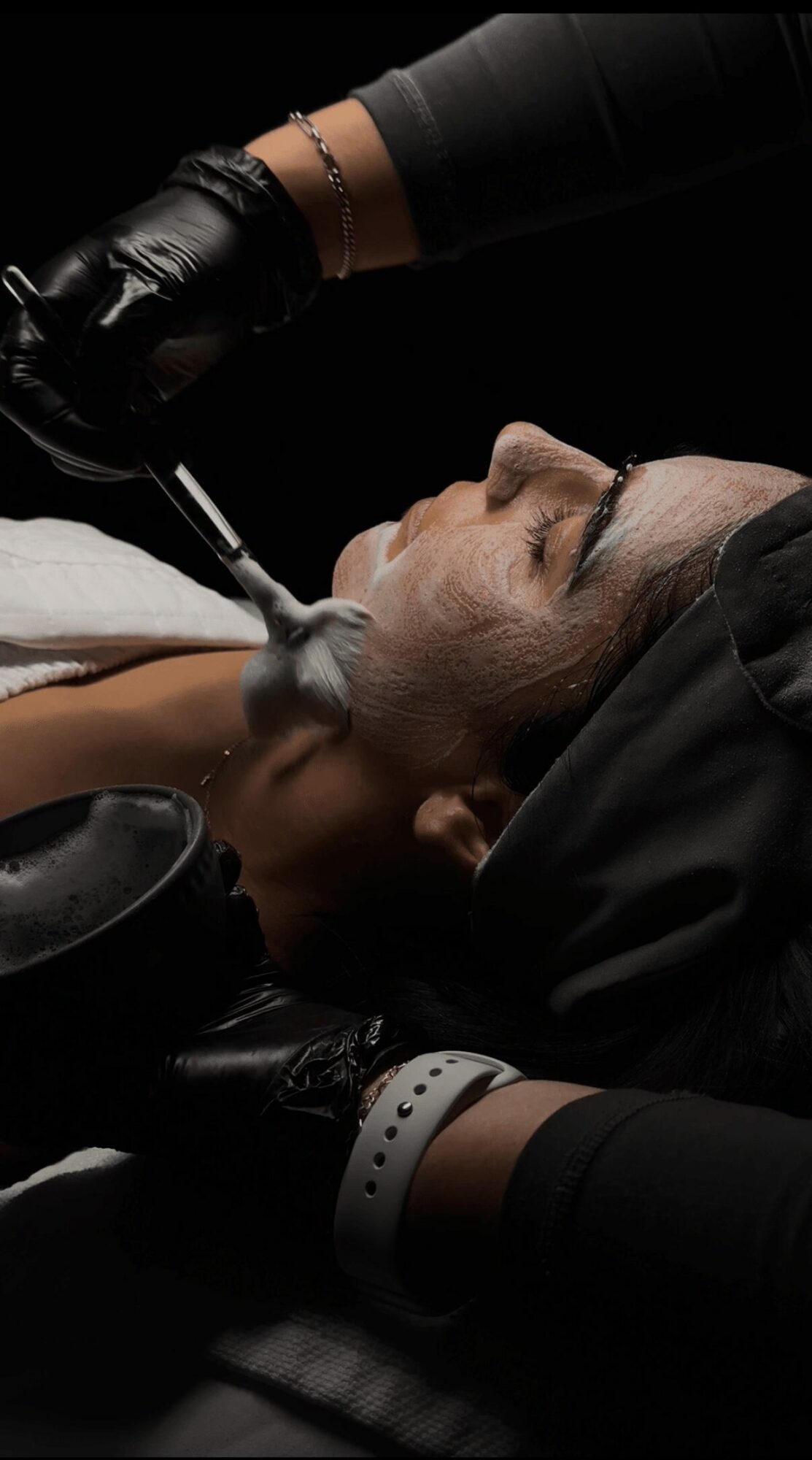 Person receiving a facial treatment with a professional applying a mask or product, lying down with eyes closed.
