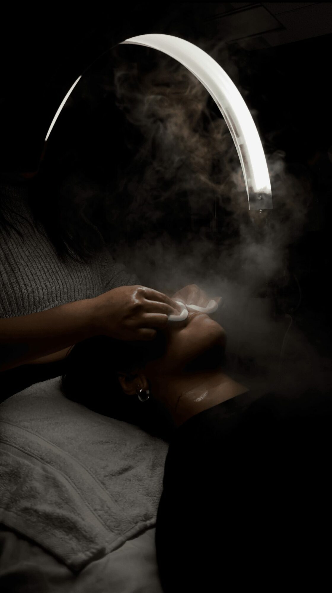 Person lying down with eyes closed, holding a lit cigarette, smoke rising, illuminated by a bright, curved light in dark surroundings.
