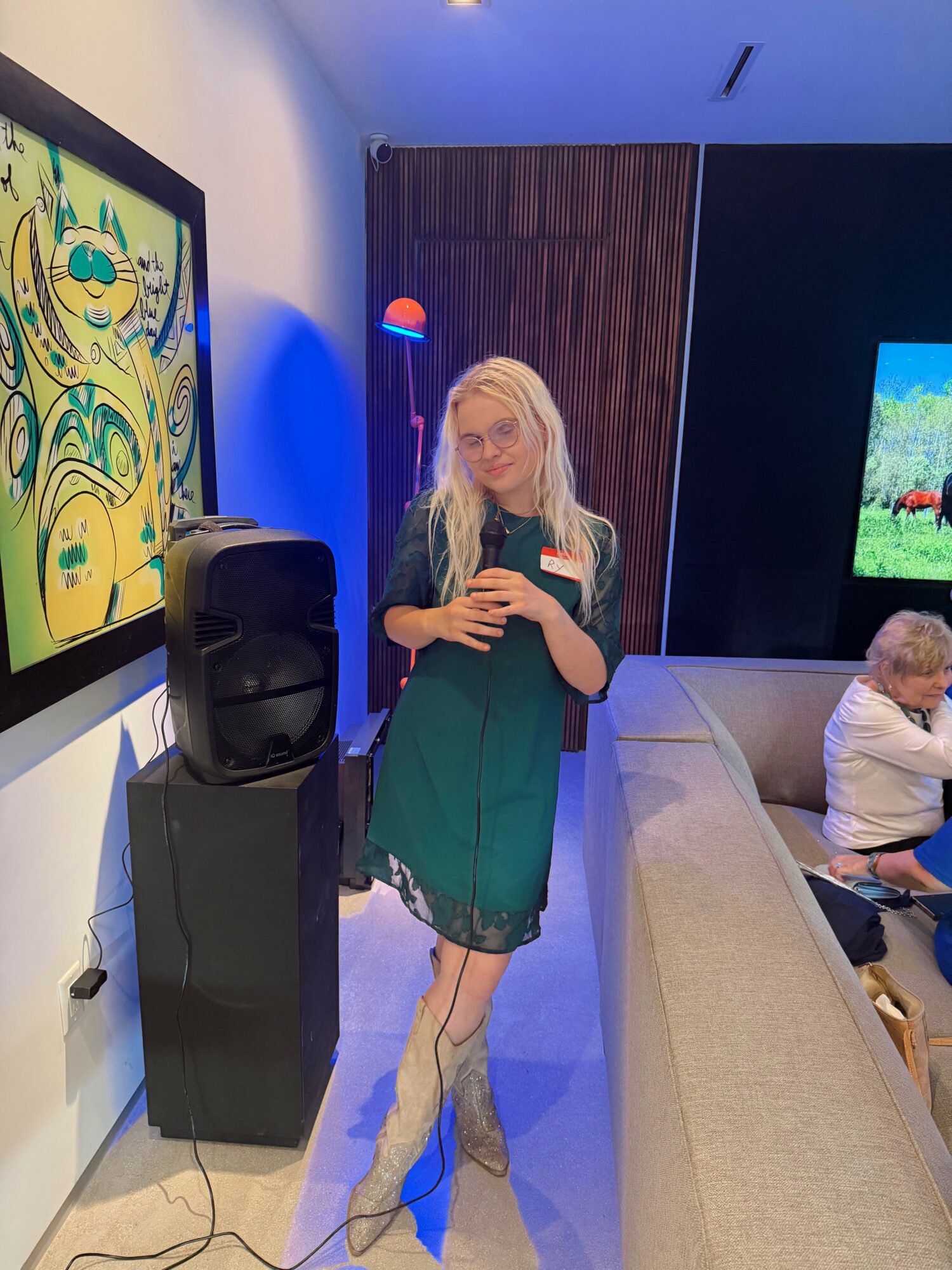 Young woman with long blonde hair taking a selfie in a room with a large screen and a painting on the wall.