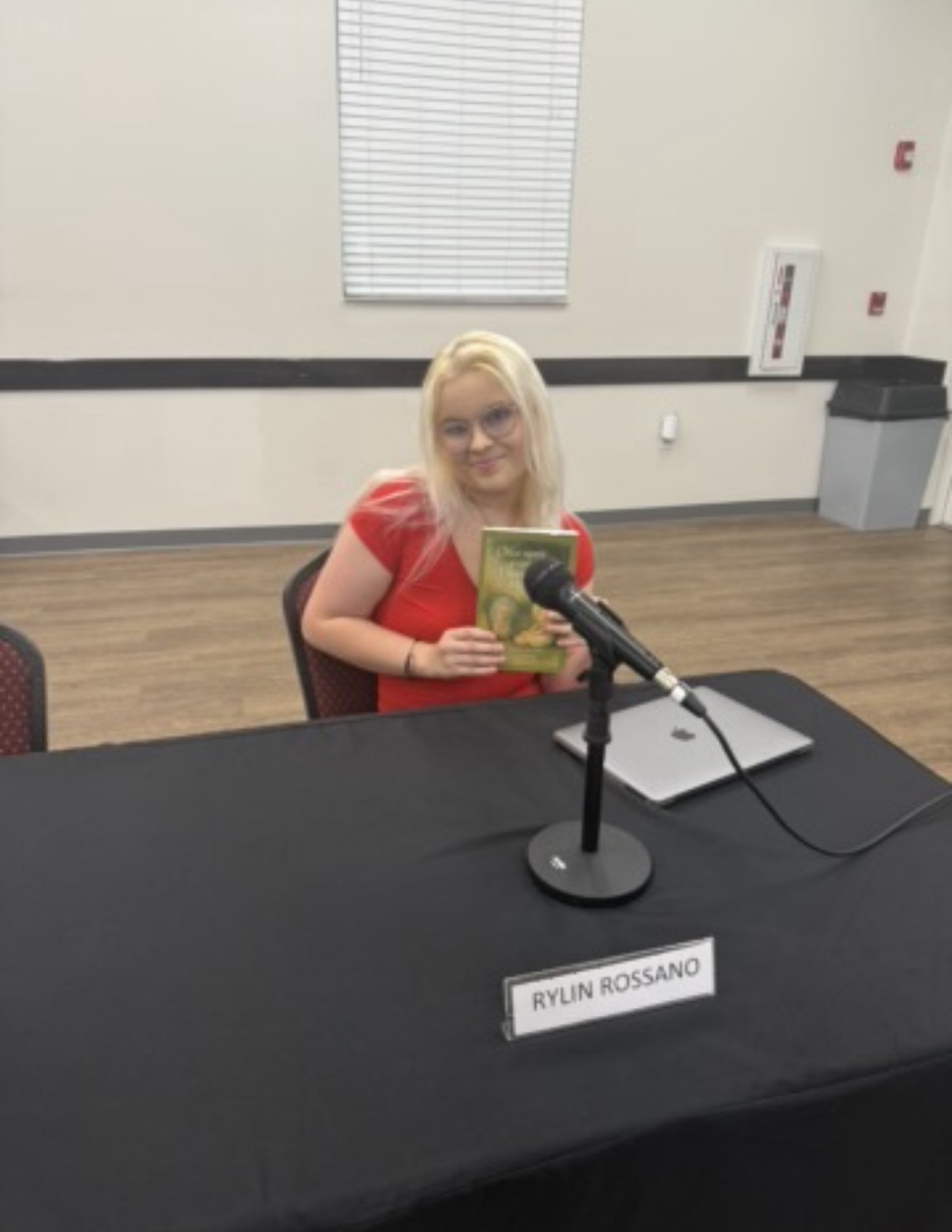Woman with blonde hair sitting at a table with a microphone, holding a book, in a room with a window and white walls.