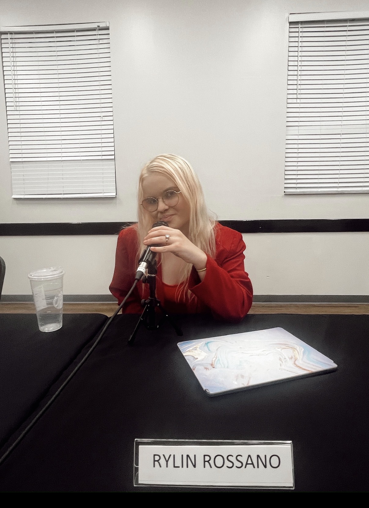 Woman with blonde hair in red sweater sitting at table with microphone, glass of water, and paper, in a room with white walls and blinds.
