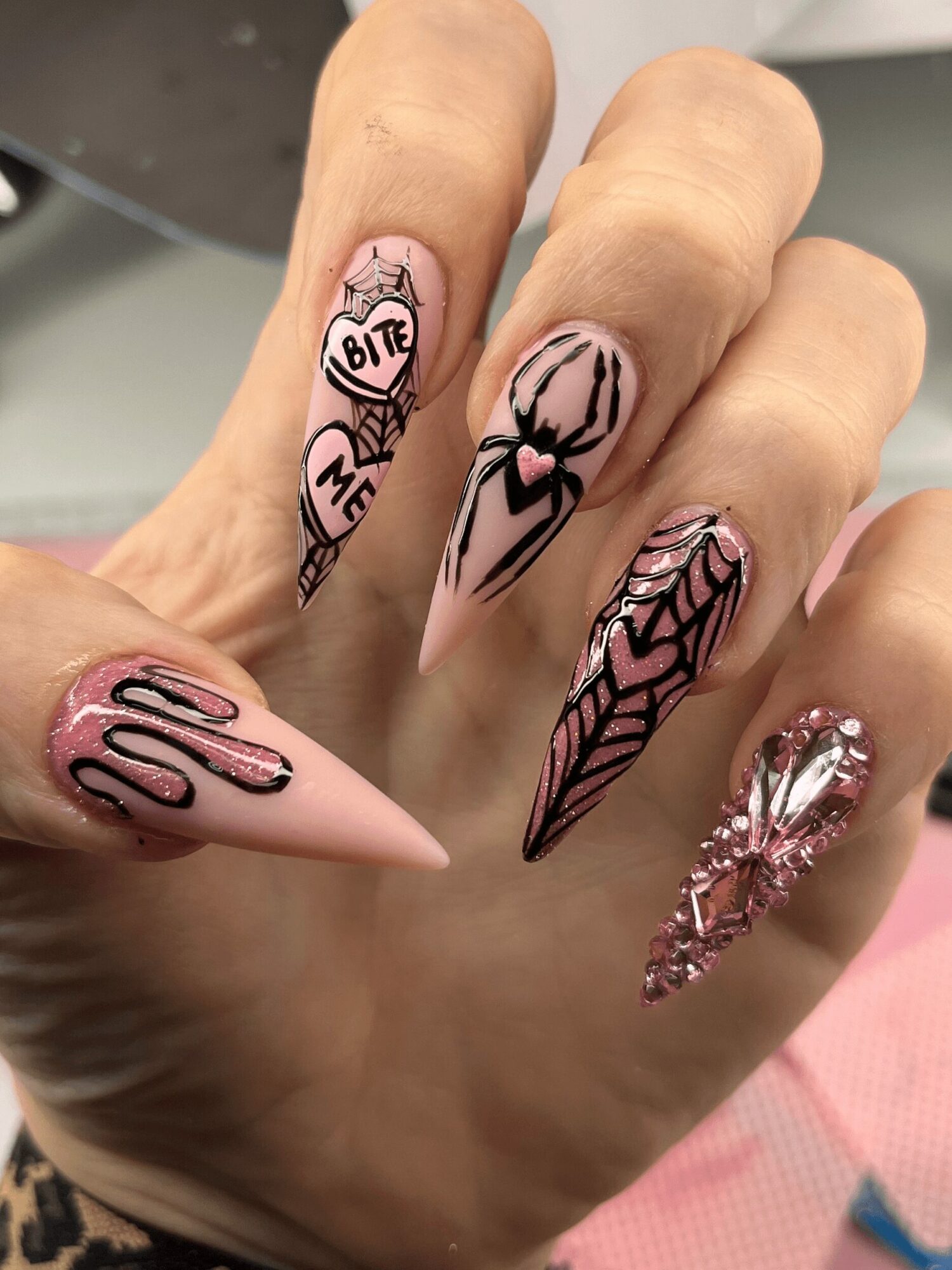 Hand with five decorated nails featuring black and pink designs, including hearts, stripes, and a spider web pattern.