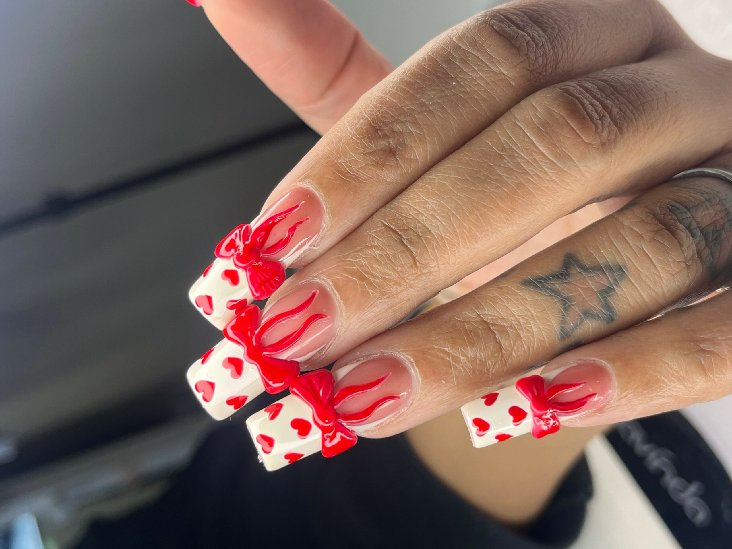 Hand with decorated nails featuring red and white heart patterns and red swirl designs, with a star tattoo visible on finger.