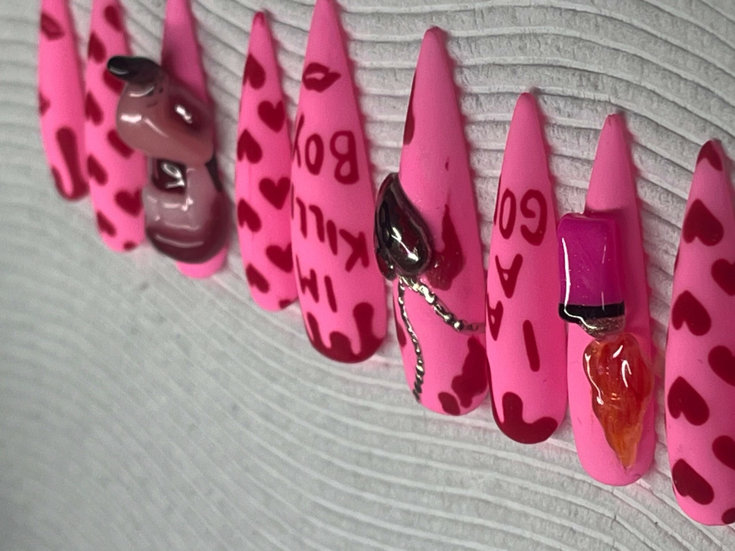 Close-up of pink nail tips with red heart patterns and various decorative elements, on a textured surface.