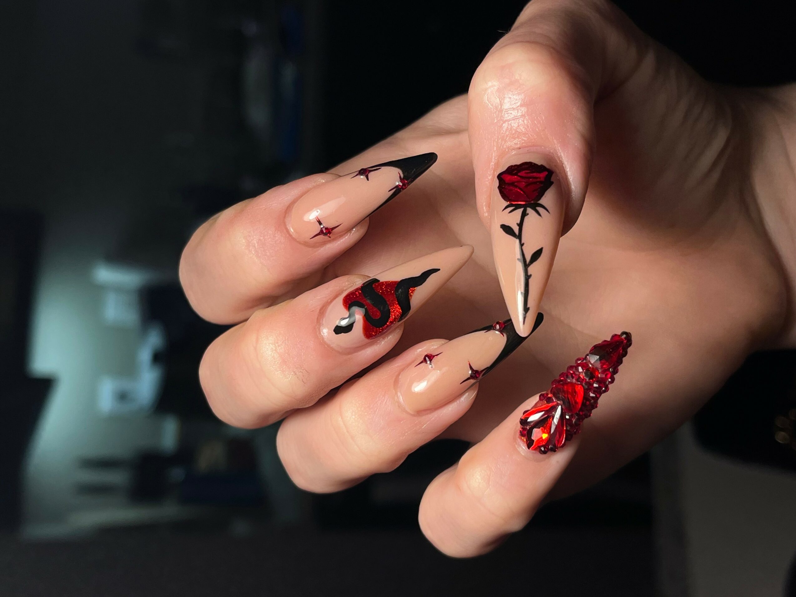 Hand with long, decorated nails featuring black, red, and floral designs, including a rose and abstract patterns.