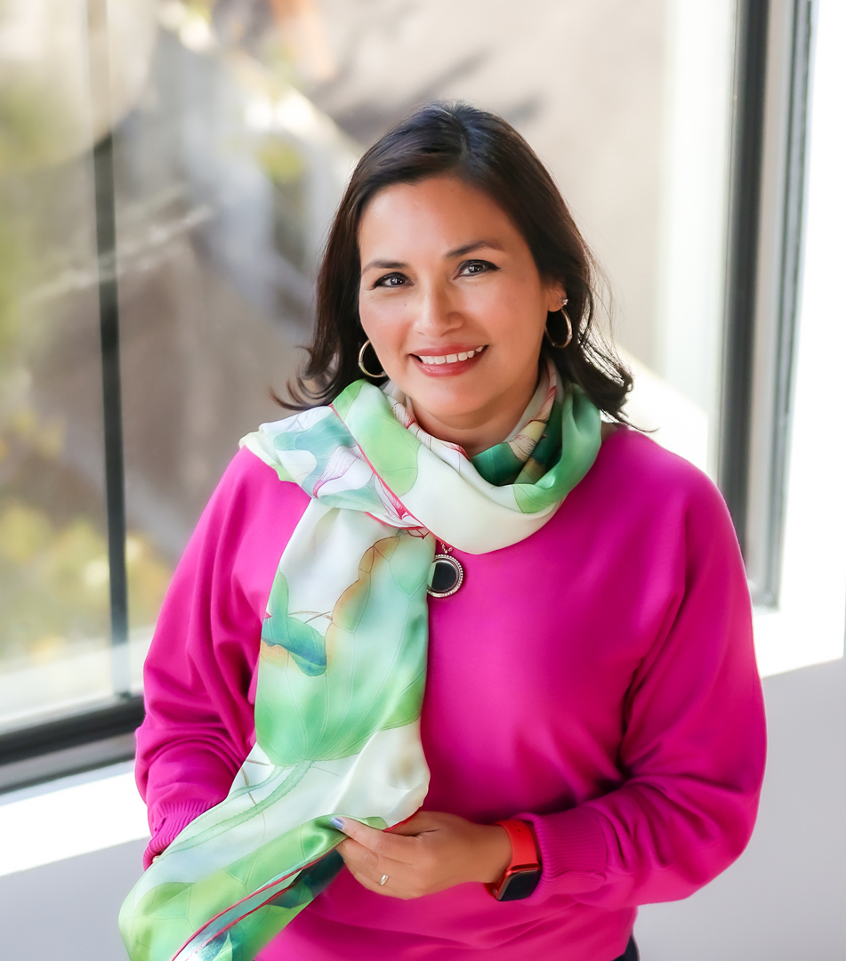 Woman with dark hair smiling, wearing a pink top and green scarf, sitting near a window.