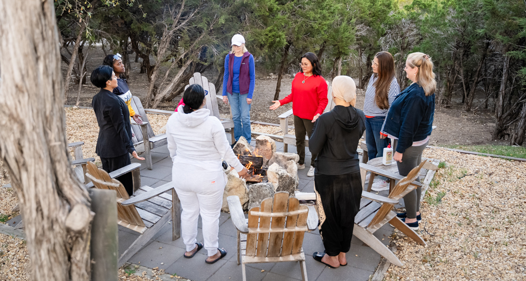 Group of people gathered outdoors around a fire pit with chairs, trees in background, some holding papers or books.