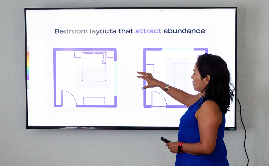 Woman in blue dress points at a presentation screen displaying bedroom layouts and text about attracting abundance.