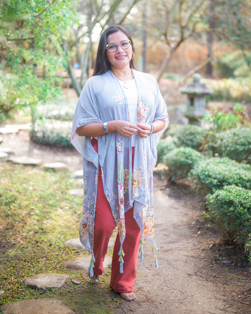 Woman standing outdoors on a garden path, smiling, wearing glasses, a light blue shawl, red pants, and a necklace.