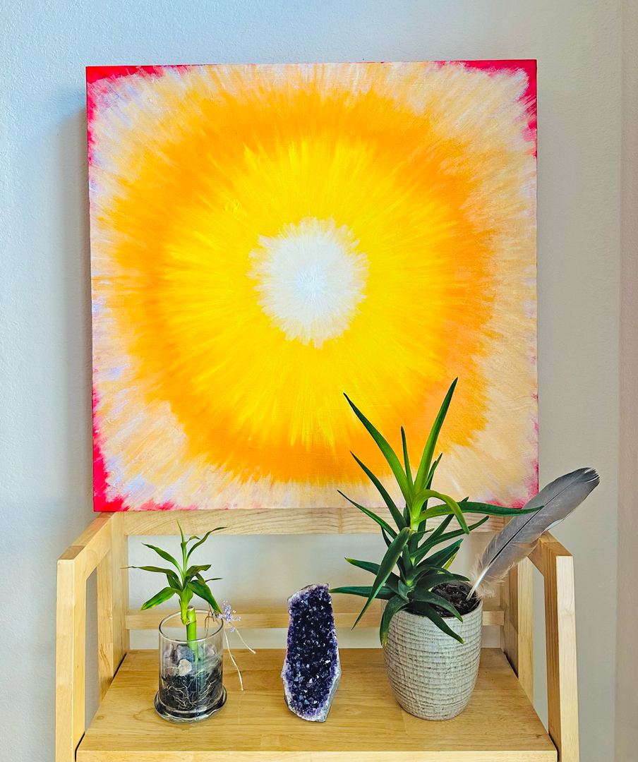Abstract painting with yellow, orange, white, and pink colors, placed on a wooden table with plants and a crystal.