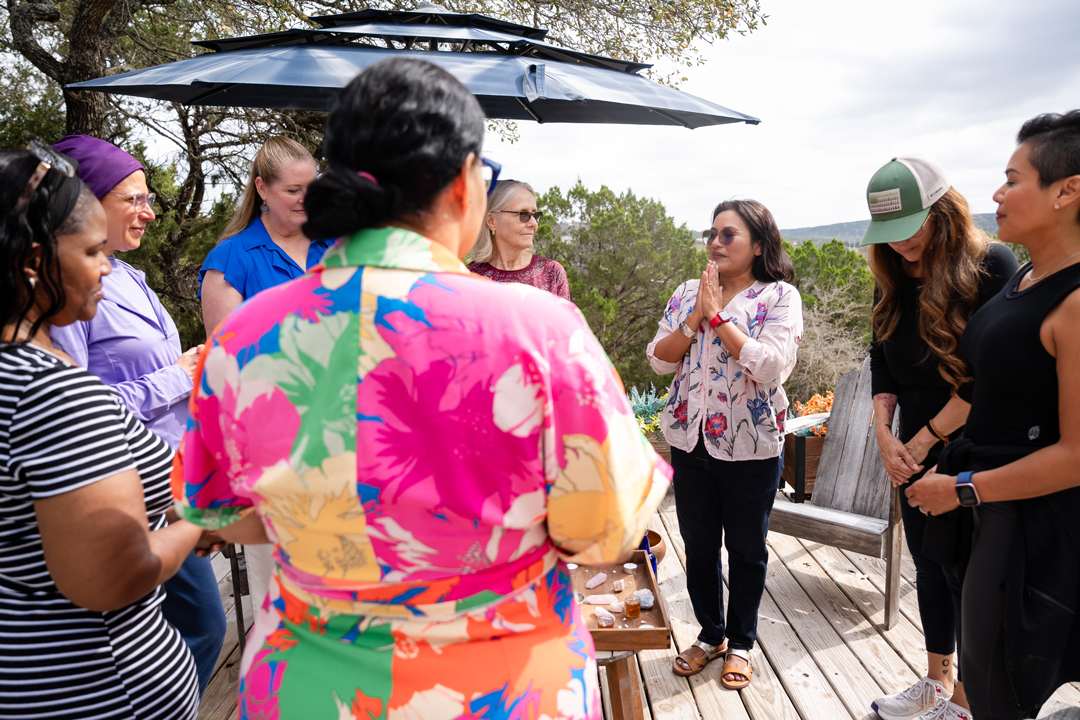 Group of women standing outdoors on a wooden deck, engaged in conversation, with trees and sky in background.