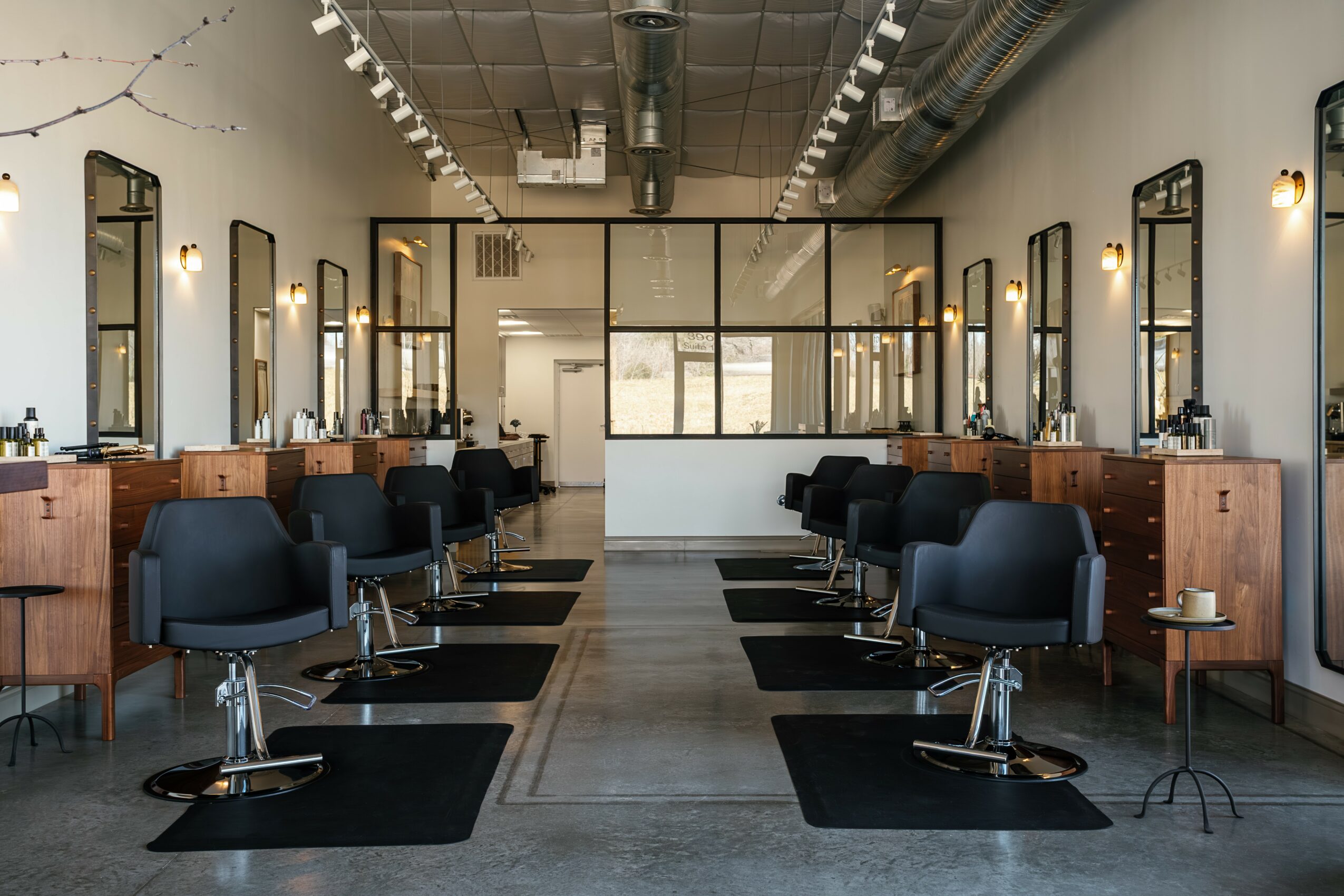 Empty hair salon with chairs, mirrors, and styling stations, illuminated by overhead lights and natural light from windows.