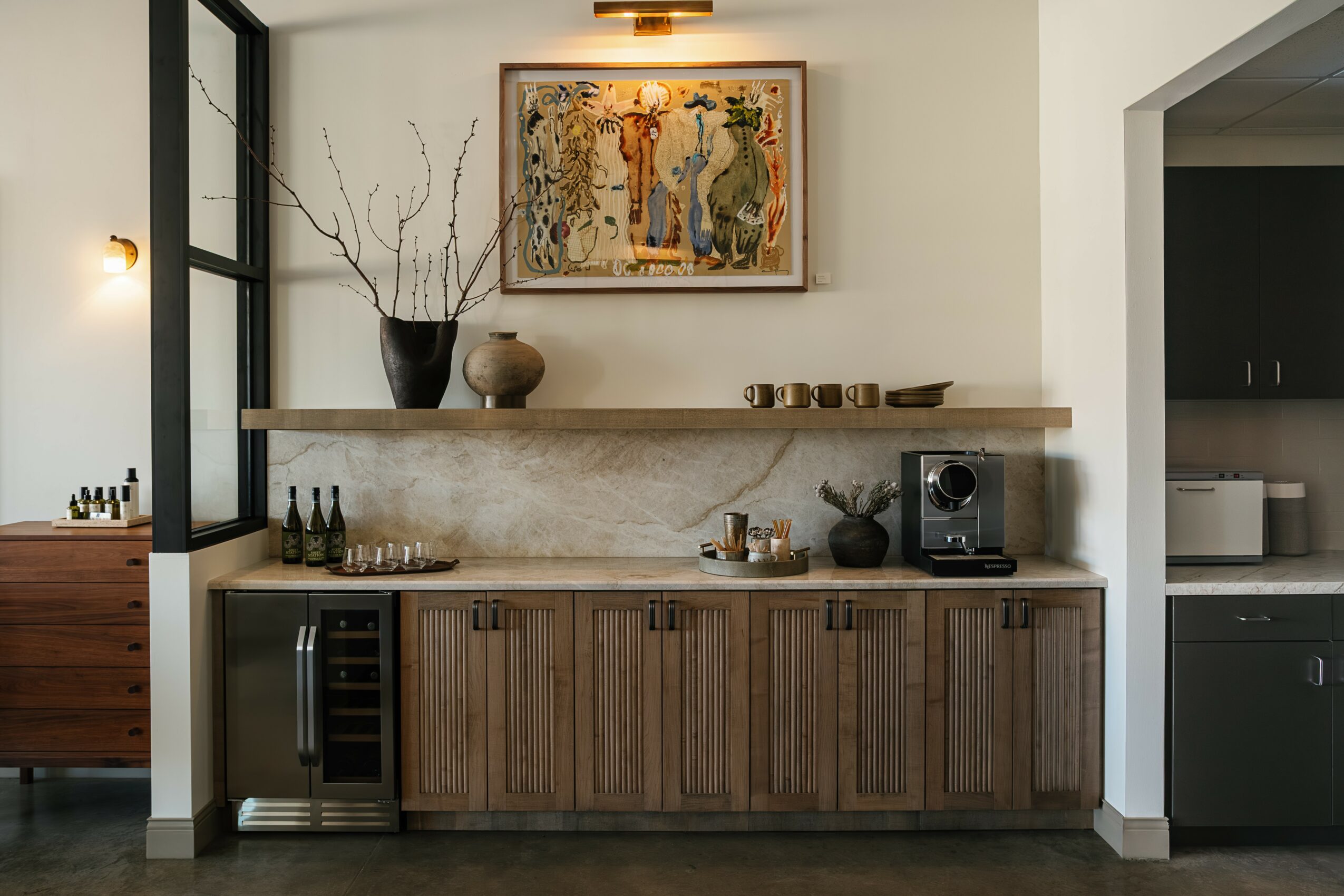 Wooden cabinet with a wine cooler, decorative items, and a small speaker, against a white wall with a shelf and artwork.