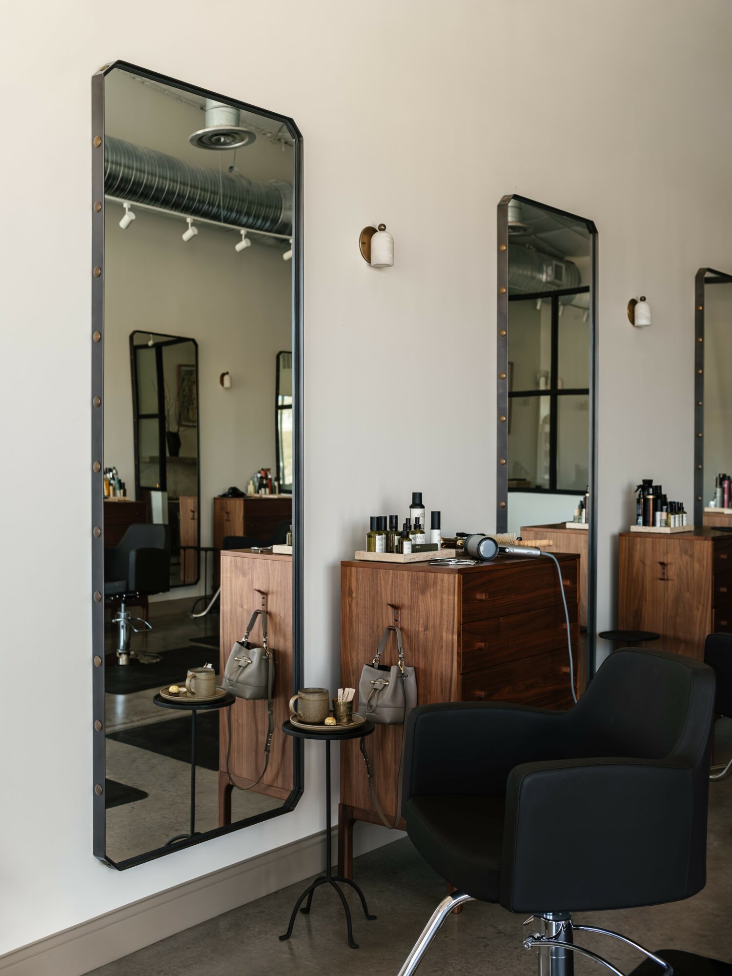 Salon with tall mirror, wooden counter, black chair, and small table with items on it, against a white wall.