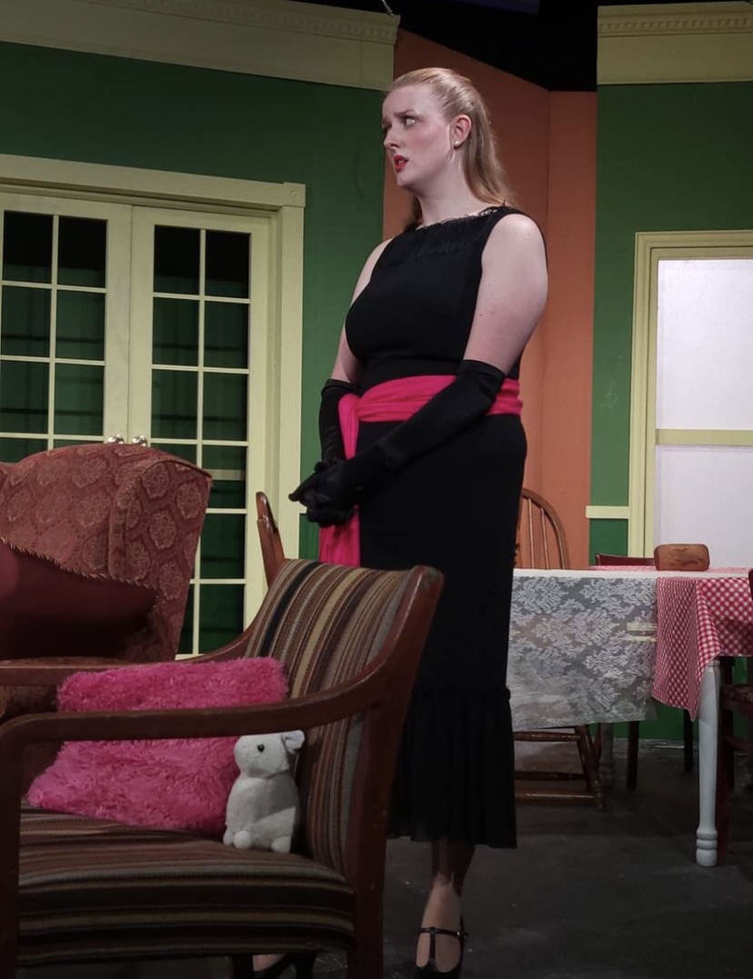 Woman in black dress with pink sash stands in room with green and yellow walls, furniture, and a table with a pink tablecloth.