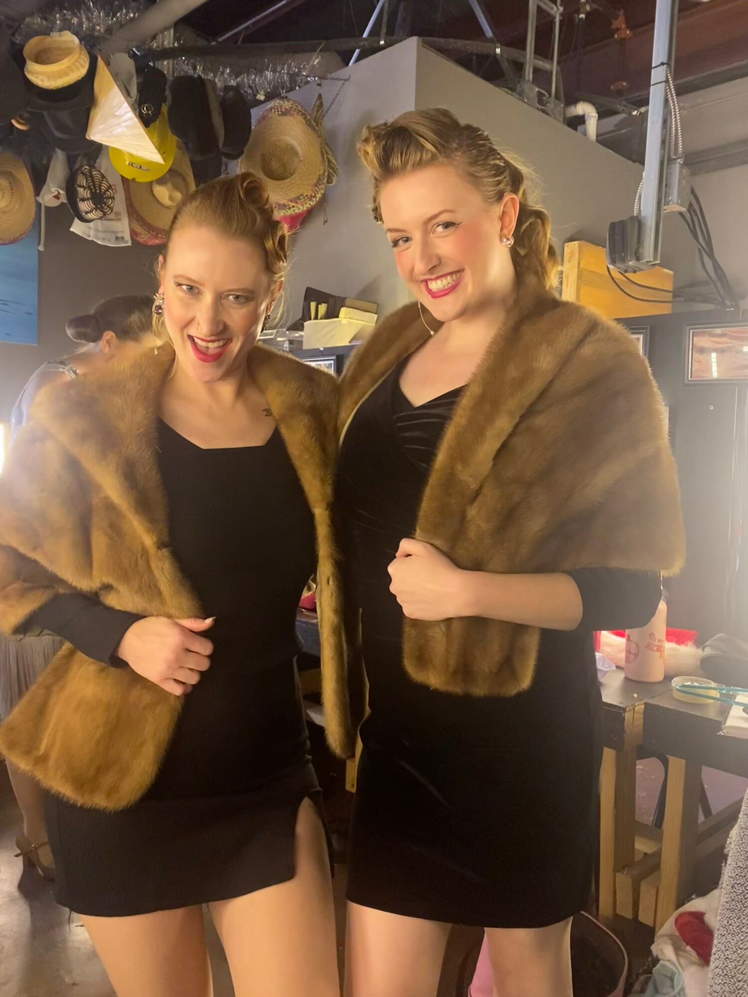 Two women in black dresses and fur stoles smiling indoors with hats and clothing in background.