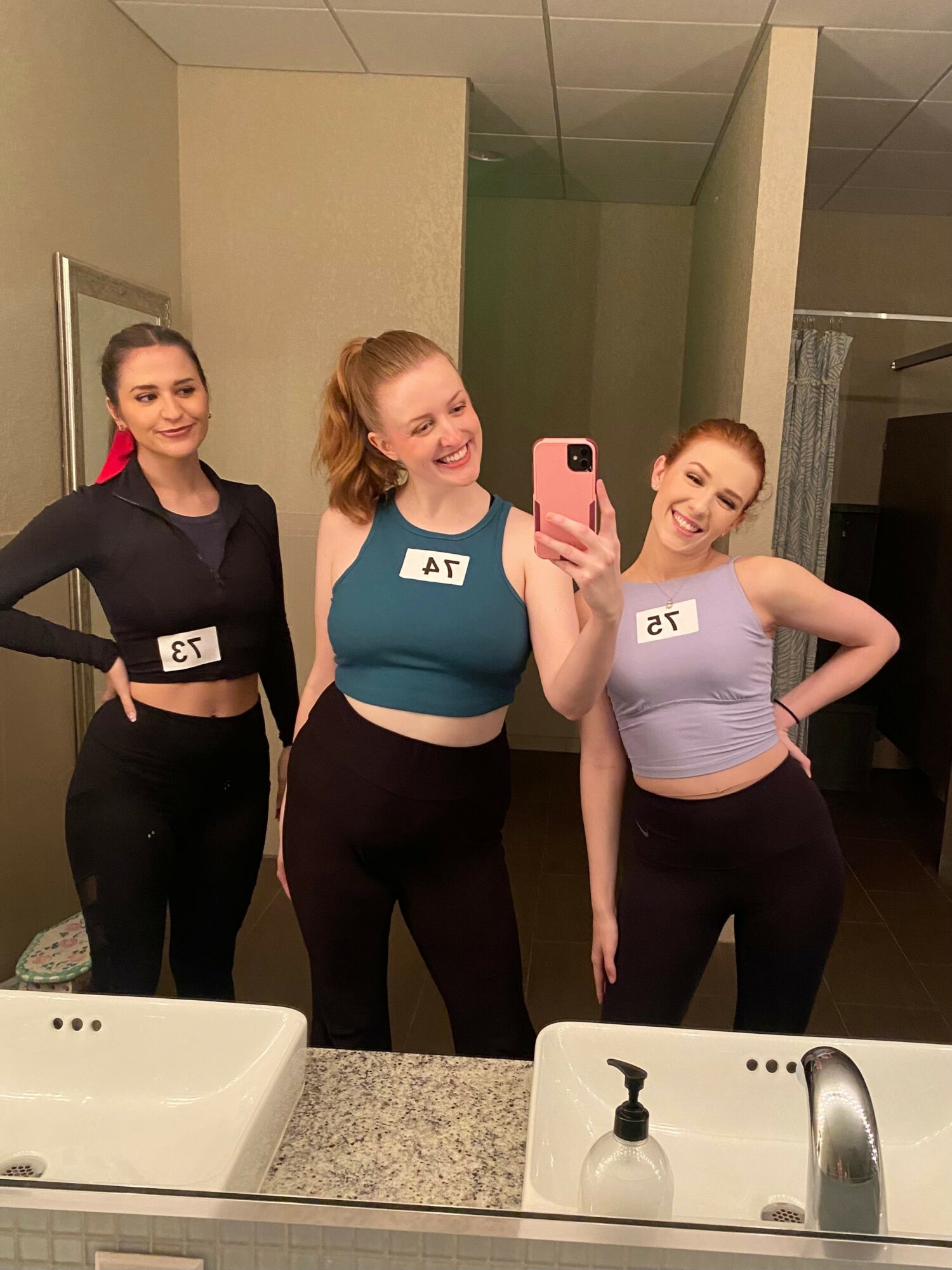 Three women taking a mirror selfie in a bathroom, smiling, wearing athletic clothing, with numbered tags on their shirts.