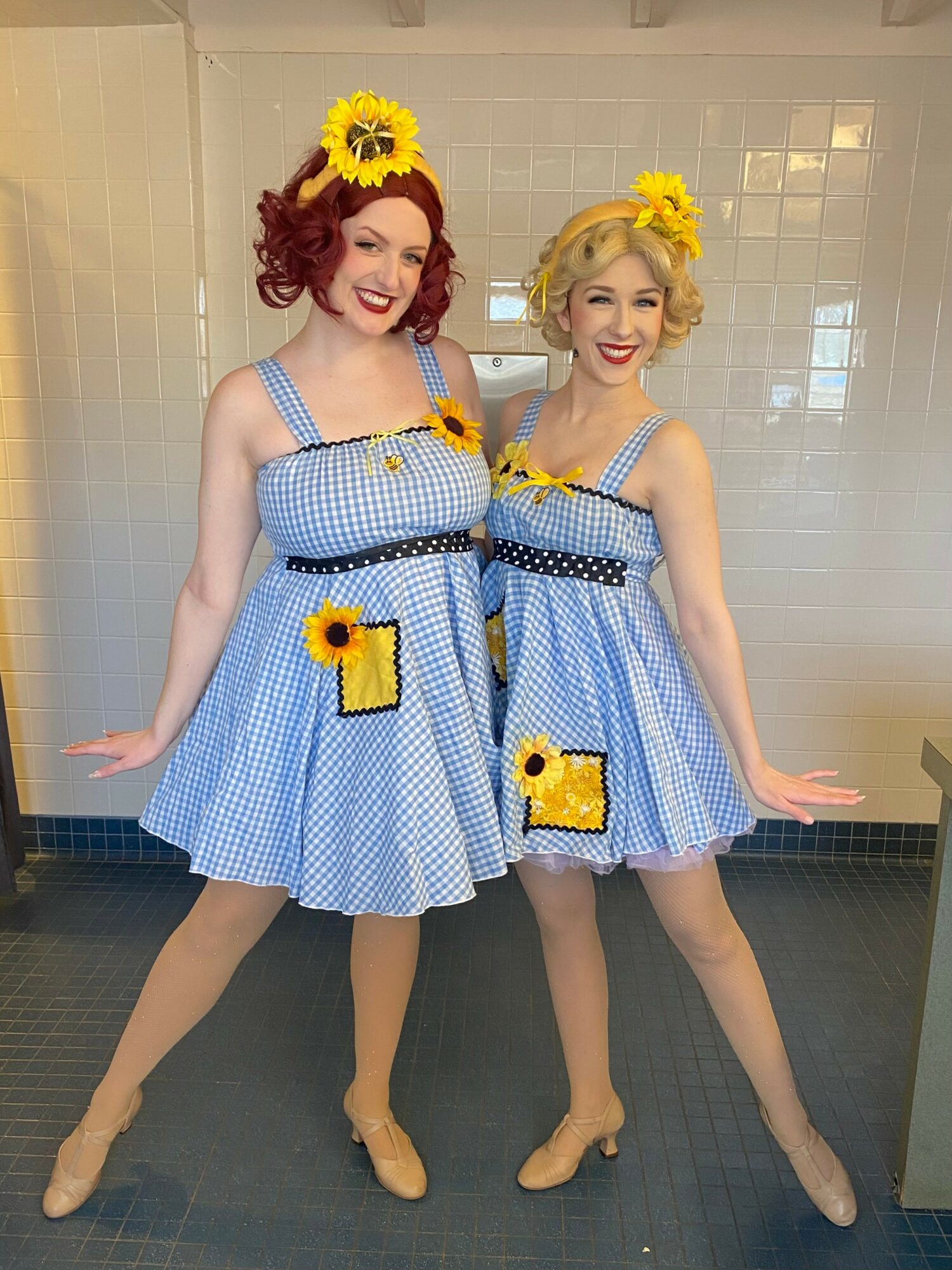 Two women in vintage-style blue gingham dresses with sunflower accessories, smiling and posing in front of a tiled wall.