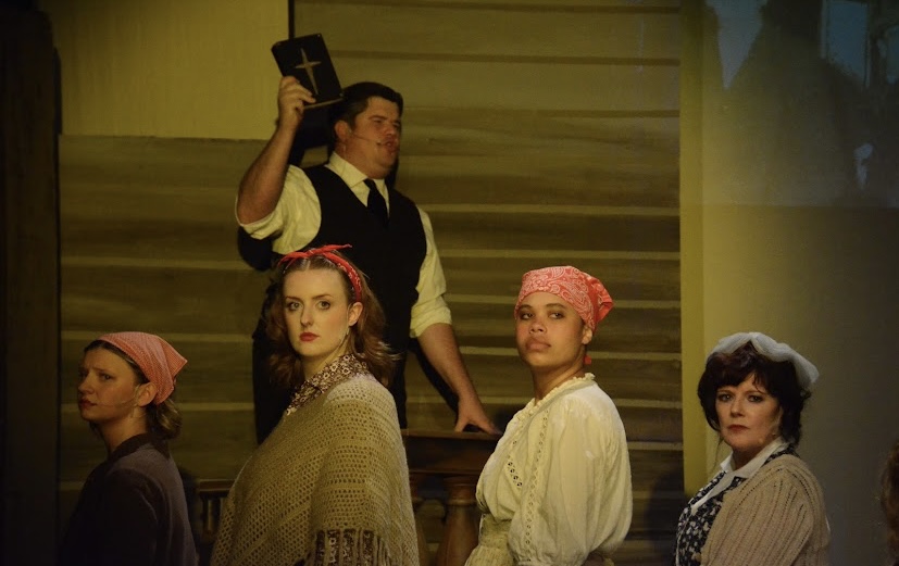 Four women and one man on stage, woman in background holding a cross, dressed in period costumes, wooden backdrop.