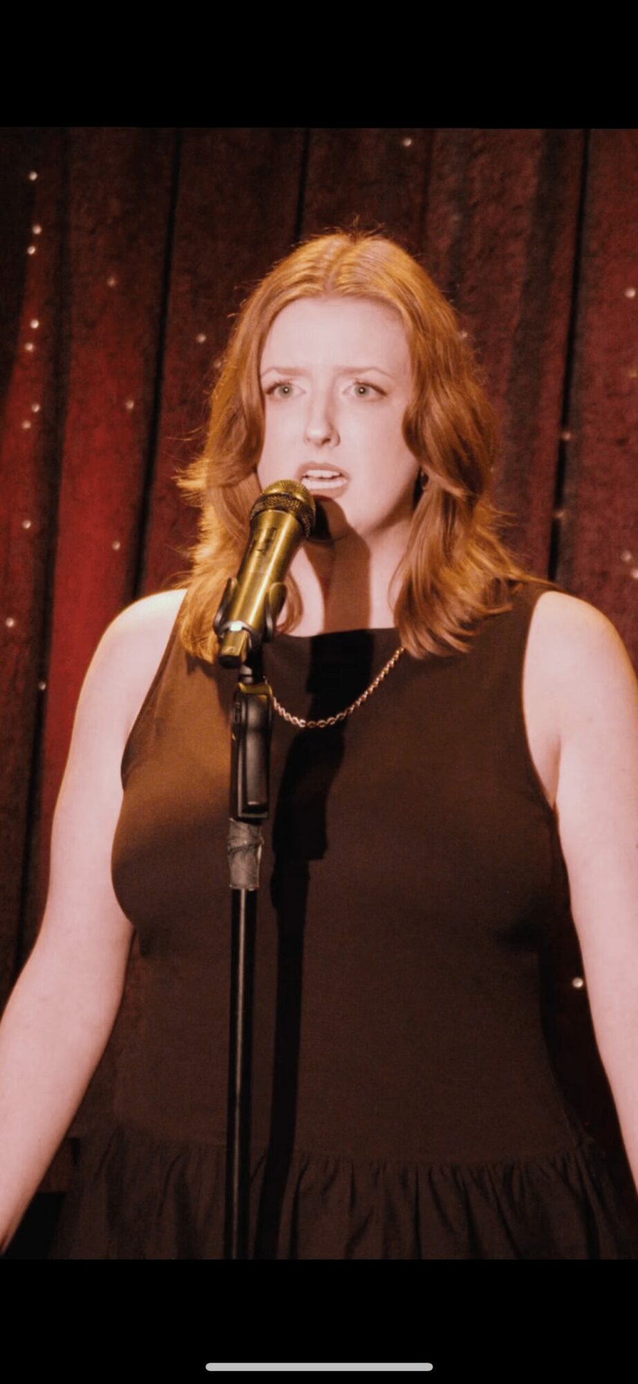 Woman with red hair singing into a gold microphone on stage with a dark curtain background.