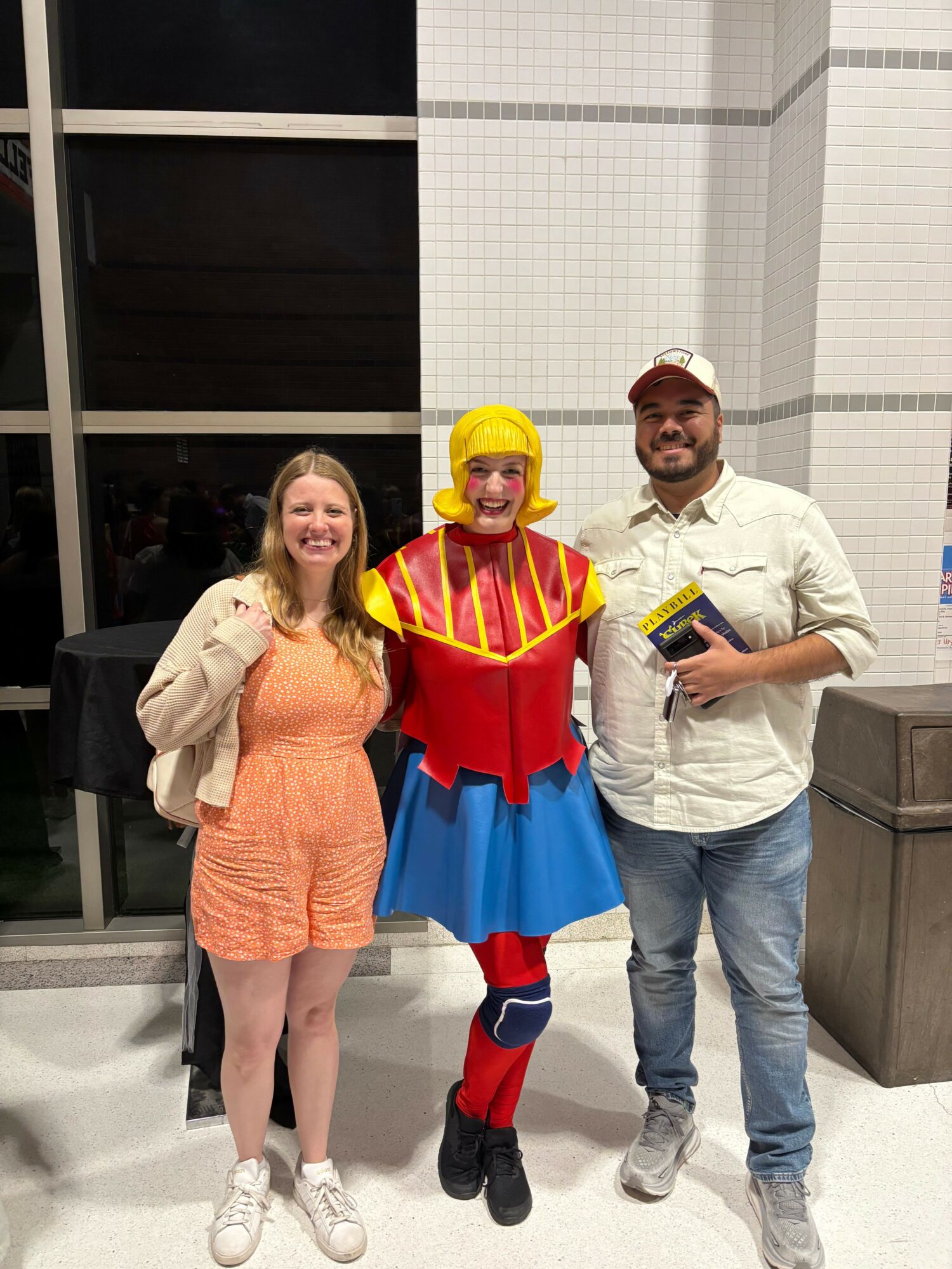 Two people standing next to a clown in a colorful costume inside a building with tiled walls.