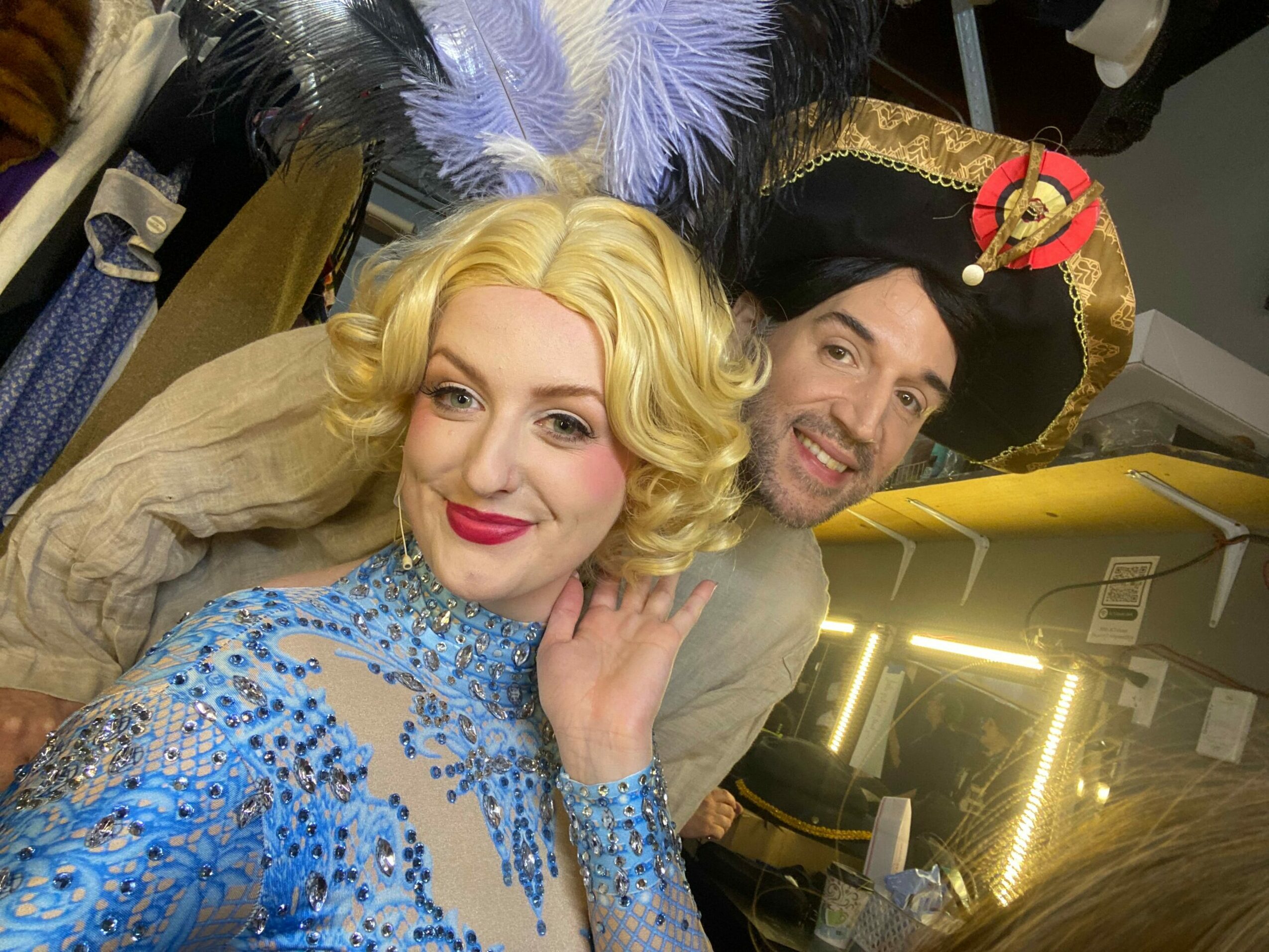 Two people dressed in costumes, one with blonde wig and makeup, the other in a black and gold outfit, smiling.