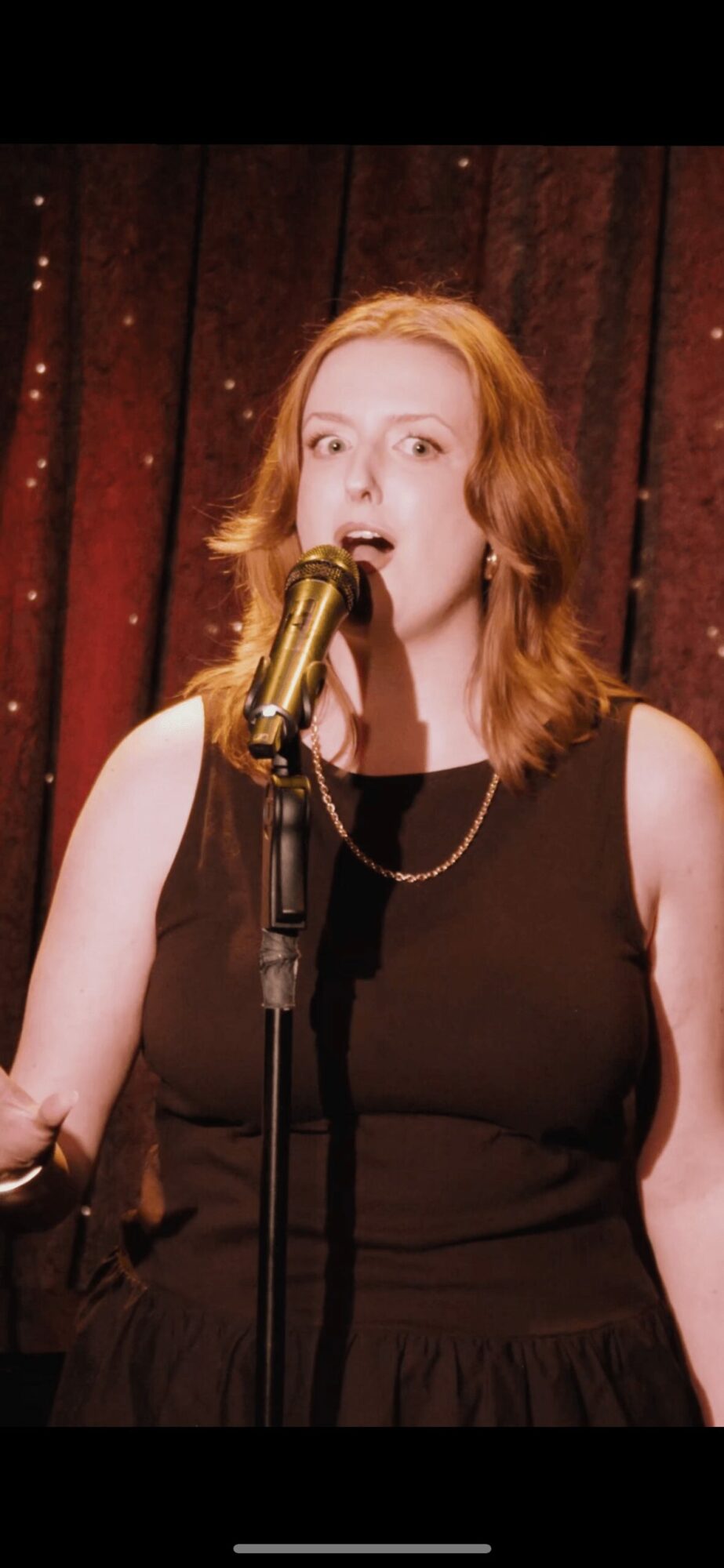 Woman with red hair singing into a microphone on stage with a dark background.