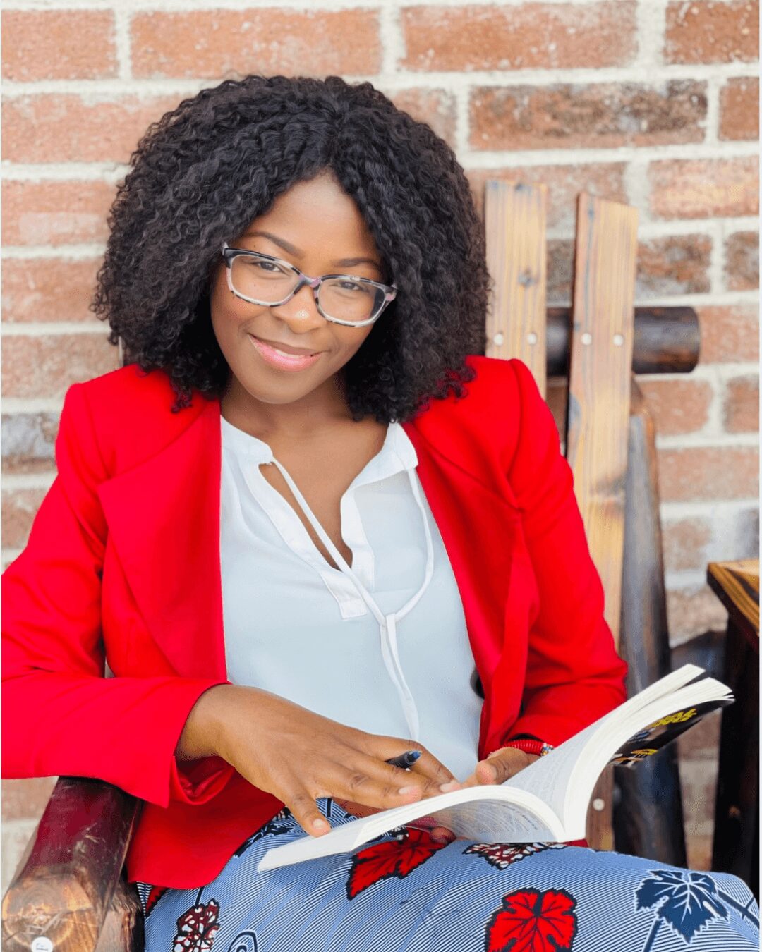 Woman with curly hair wearing glasses, red blazer, and white blouse, sitting outdoors with a brick wall background, reading a book.