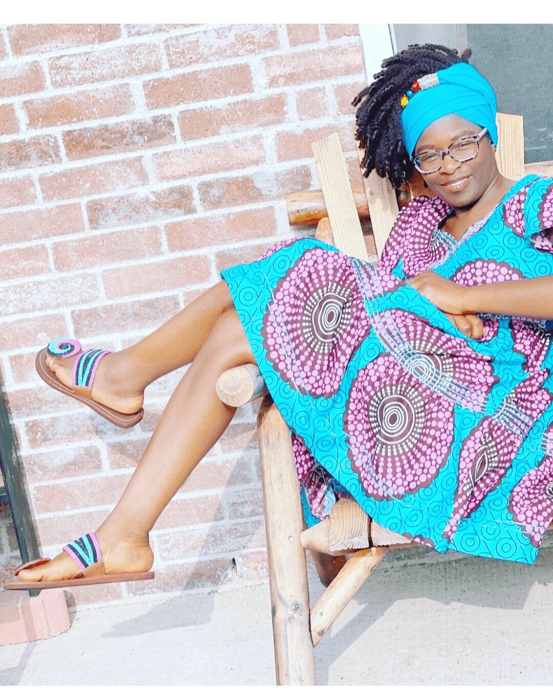 Girl with glasses and headwrap sitting on a wooden chair outdoors, wearing a colorful dress and sandals, smiling.