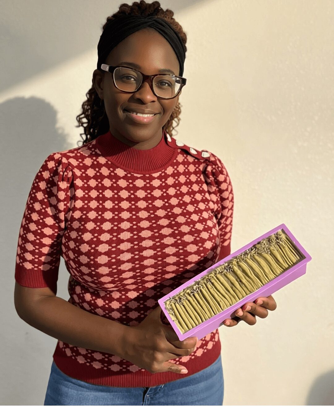 Woman with glasses and braided hair holding a purple box of yellow flowers, smiling, standing against a plain wall.