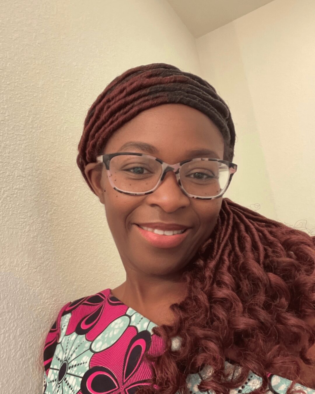 Woman with glasses and braided hair smiling, wearing a colorful patterned top, against a plain wall.