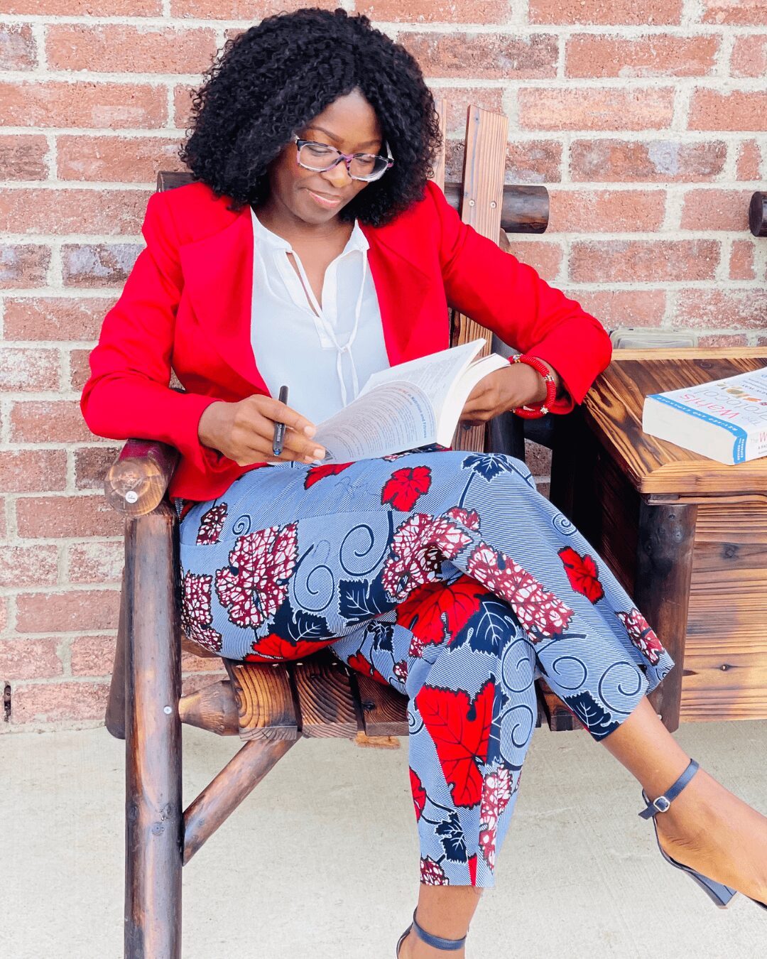 Woman with curly hair and glasses reading a book while sitting on a wooden chair against a brick wall.