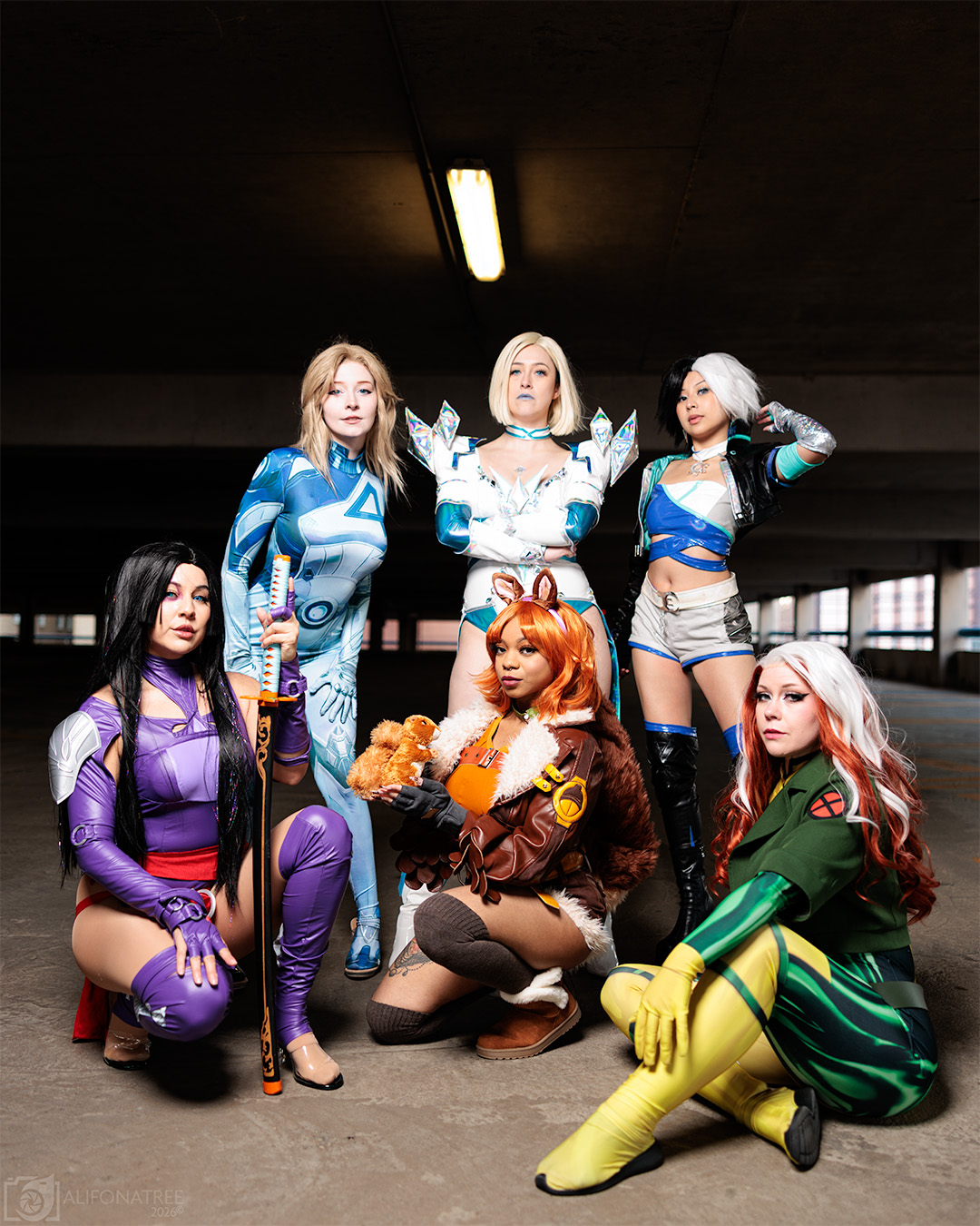 Six women in colorful costumes pose in an underground parking garage with dark ceiling and fluorescent light.