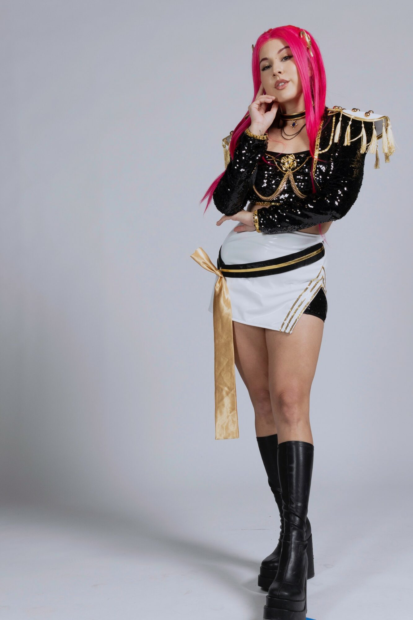 Young woman with bright pink hair in a black and white outfit with gold accents, standing with arms crossed.