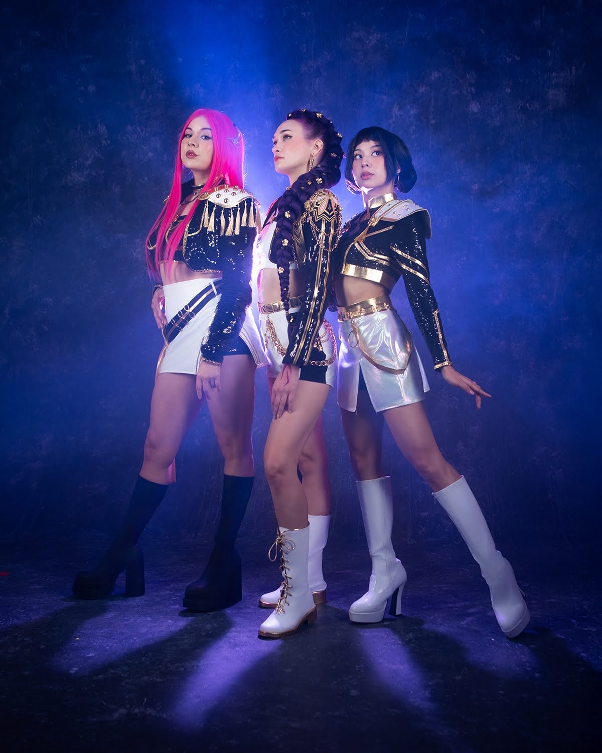 Three women in futuristic outfits stand on a dark stage with blue lighting and mist.