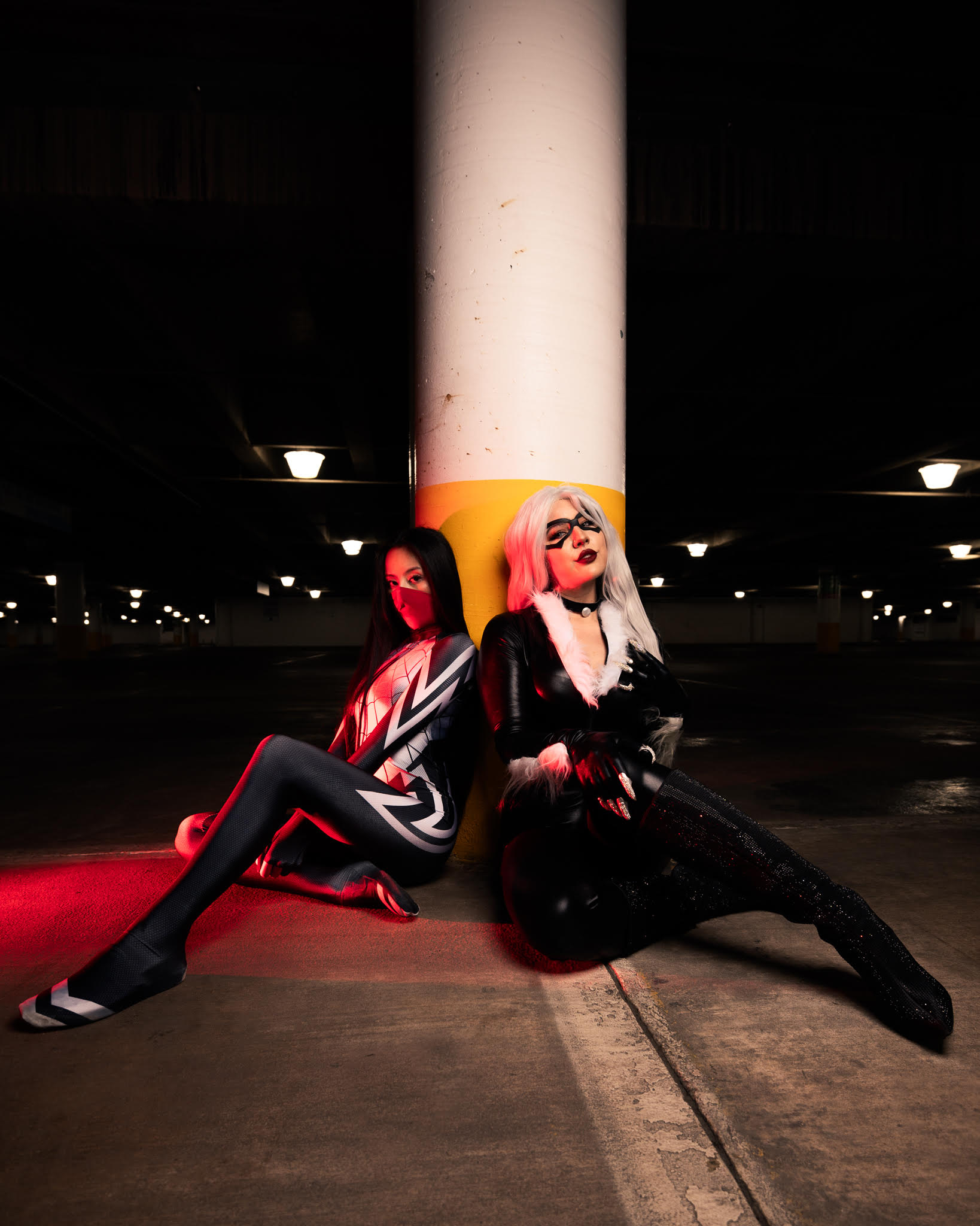 Two women with colorful hair and bold outfits sit on the ground against a large white pillar in a parking garage.