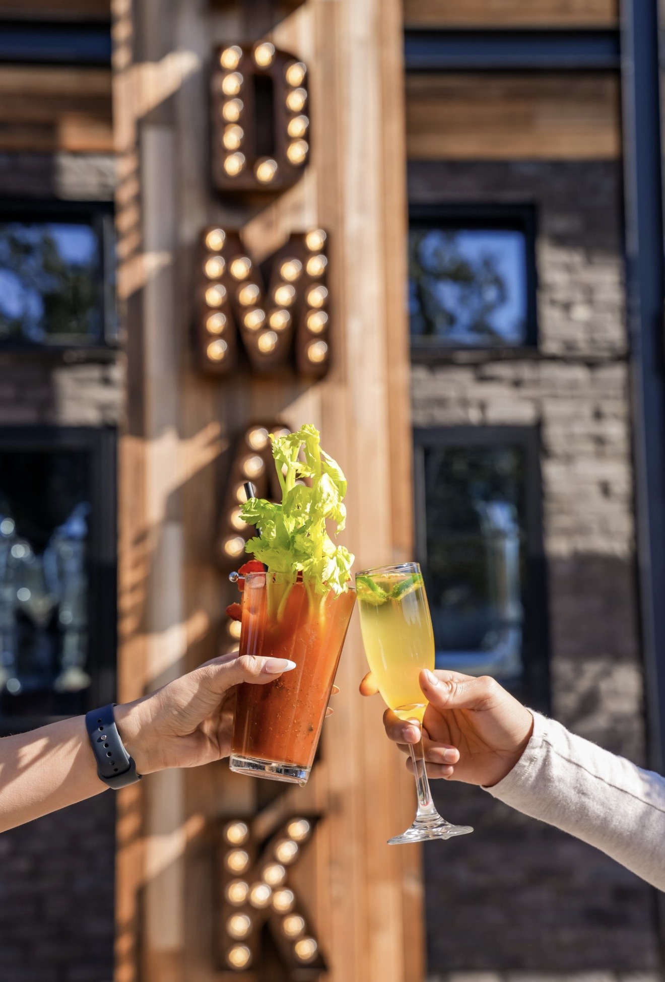 Two people hold drinks in front of a building with a vertical sign spelling 'DRINK' with illuminated bulbs.