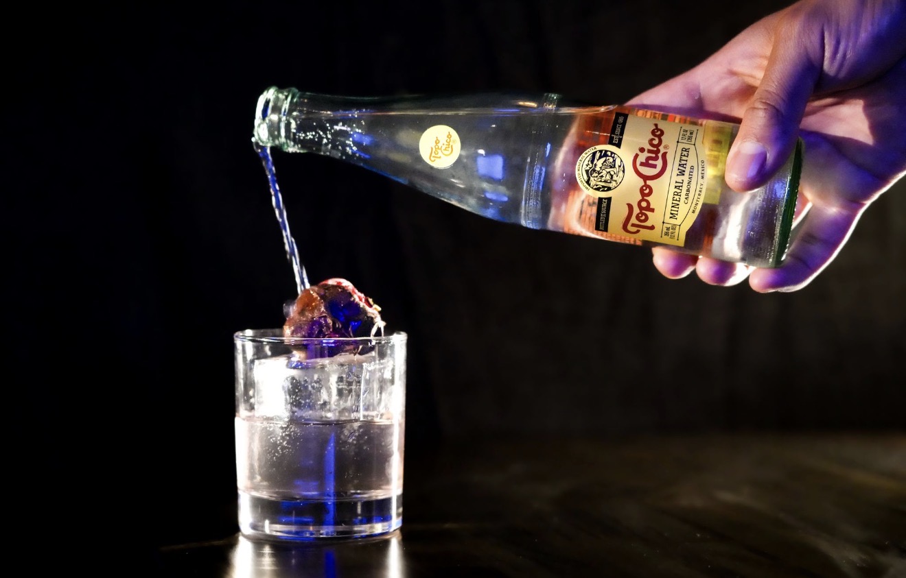Hand pouring clear liquid from a bottle into a glass with ice, dark background.