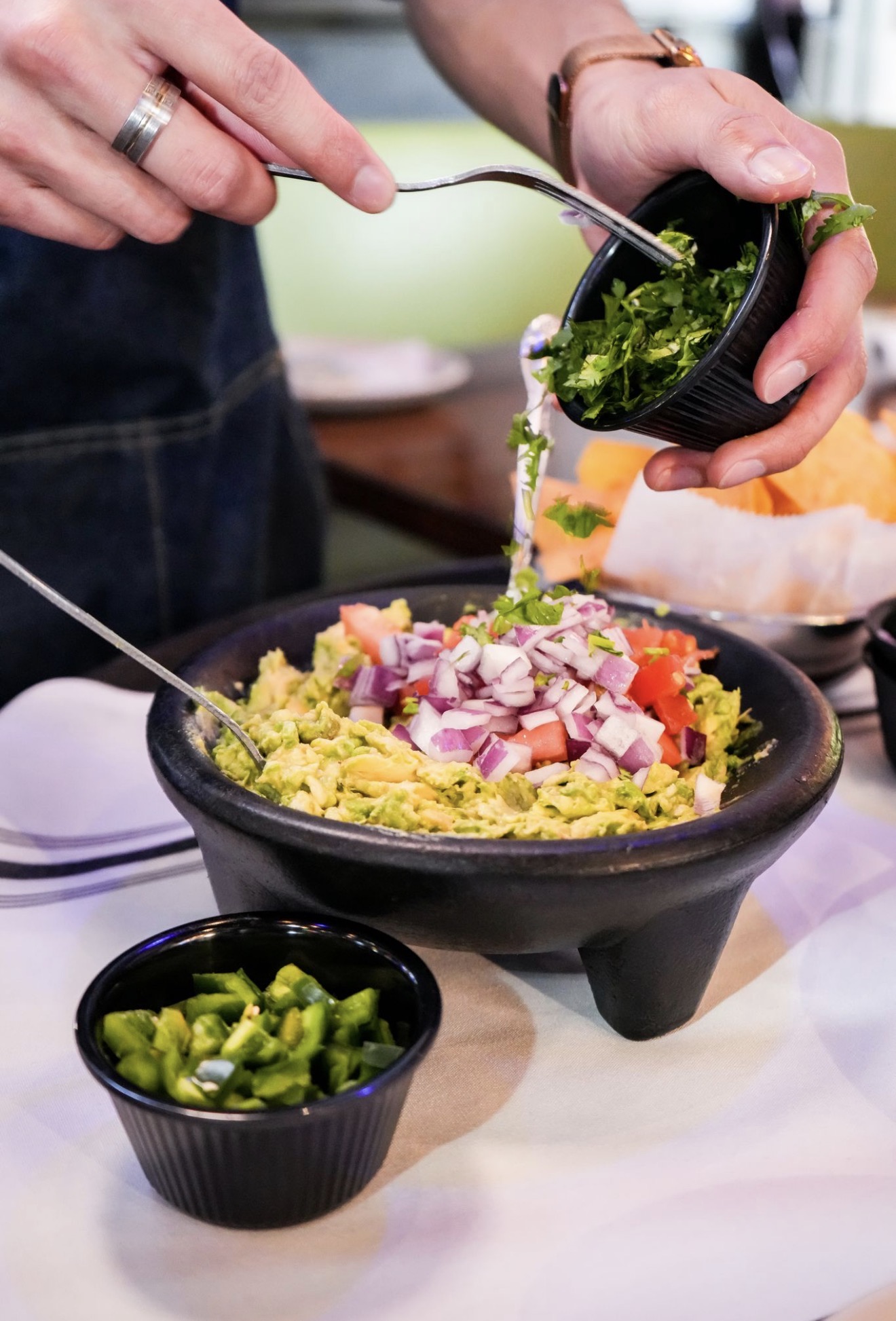 Person adding chopped herbs to a bowl of salad with diced vegetables, with a small bowl of chopped green peppers nearby.