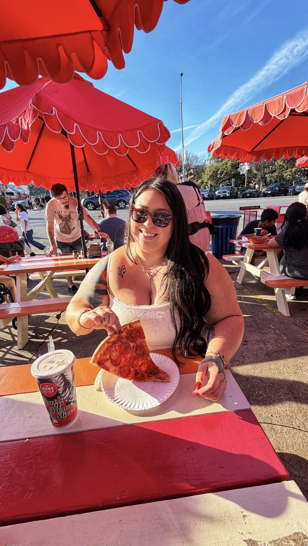 Woman with sunglasses sitting at a table outdoors, holding a slice of pizza, under red umbrellas.