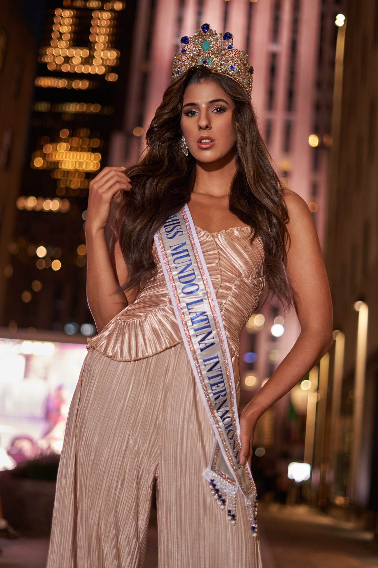 Young woman wearing a crown and sash, standing outdoors at night in an urban setting.