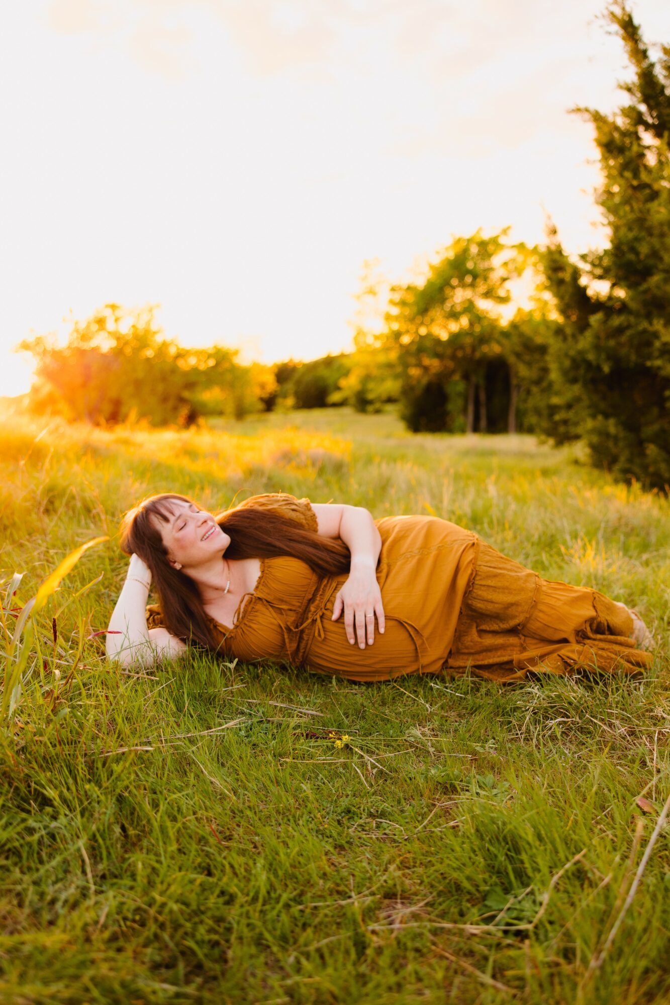 Woman lying on grass in a field during sunset, wearing a brown dress, with trees in the background.