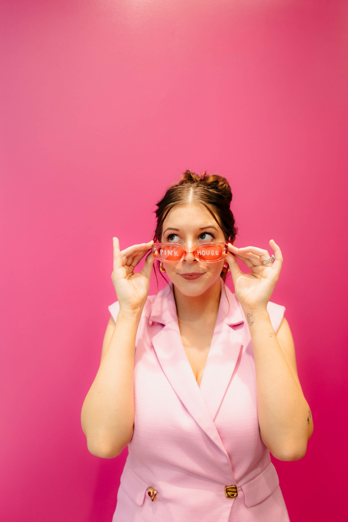 Young woman with glasses and a pink outfit holding glasses near her face, smiling, against a pink background.