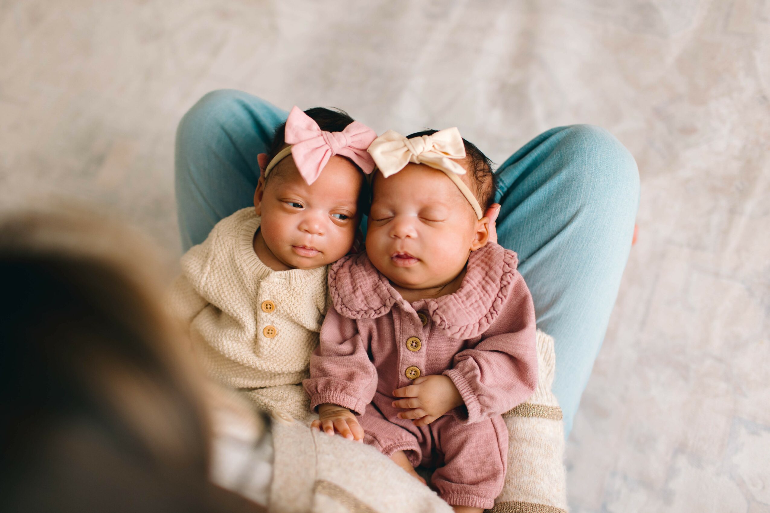 Two babies sitting on a person's lap, close together, with bows in their hair, looking relaxed and peaceful.