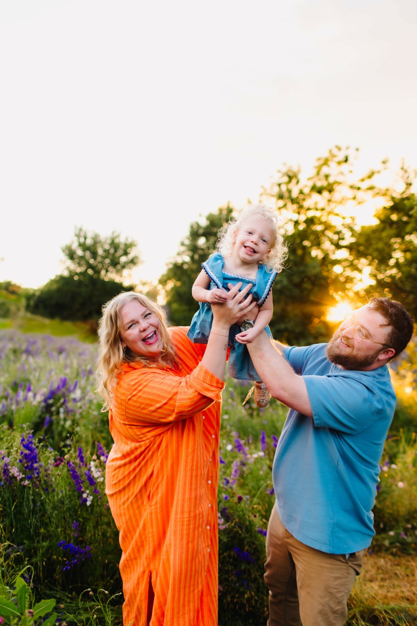 Family outdoors in a field of purple flowers, with a woman lifting a smiling child, man supporting child, sunset in background.