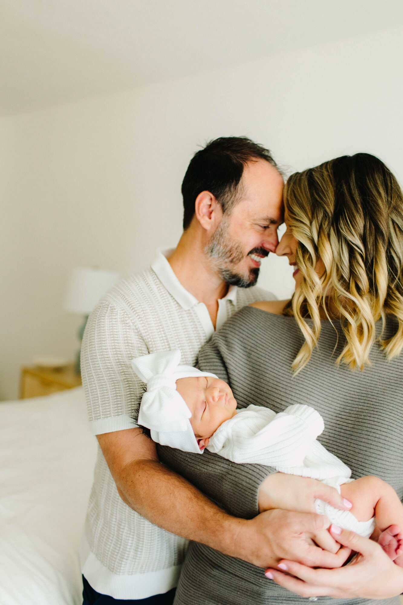 Family of three with a man, woman, and baby in a room with white walls and bed.