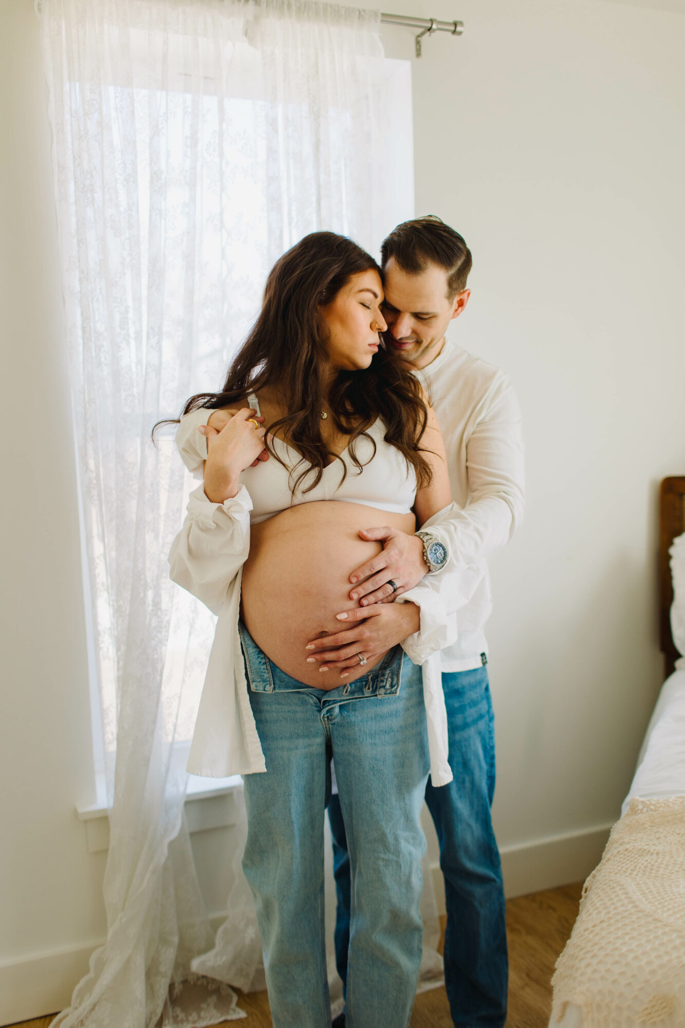 Pregnant woman and man standing together in a room, woman touching her belly, man holding her waist, near a window.
